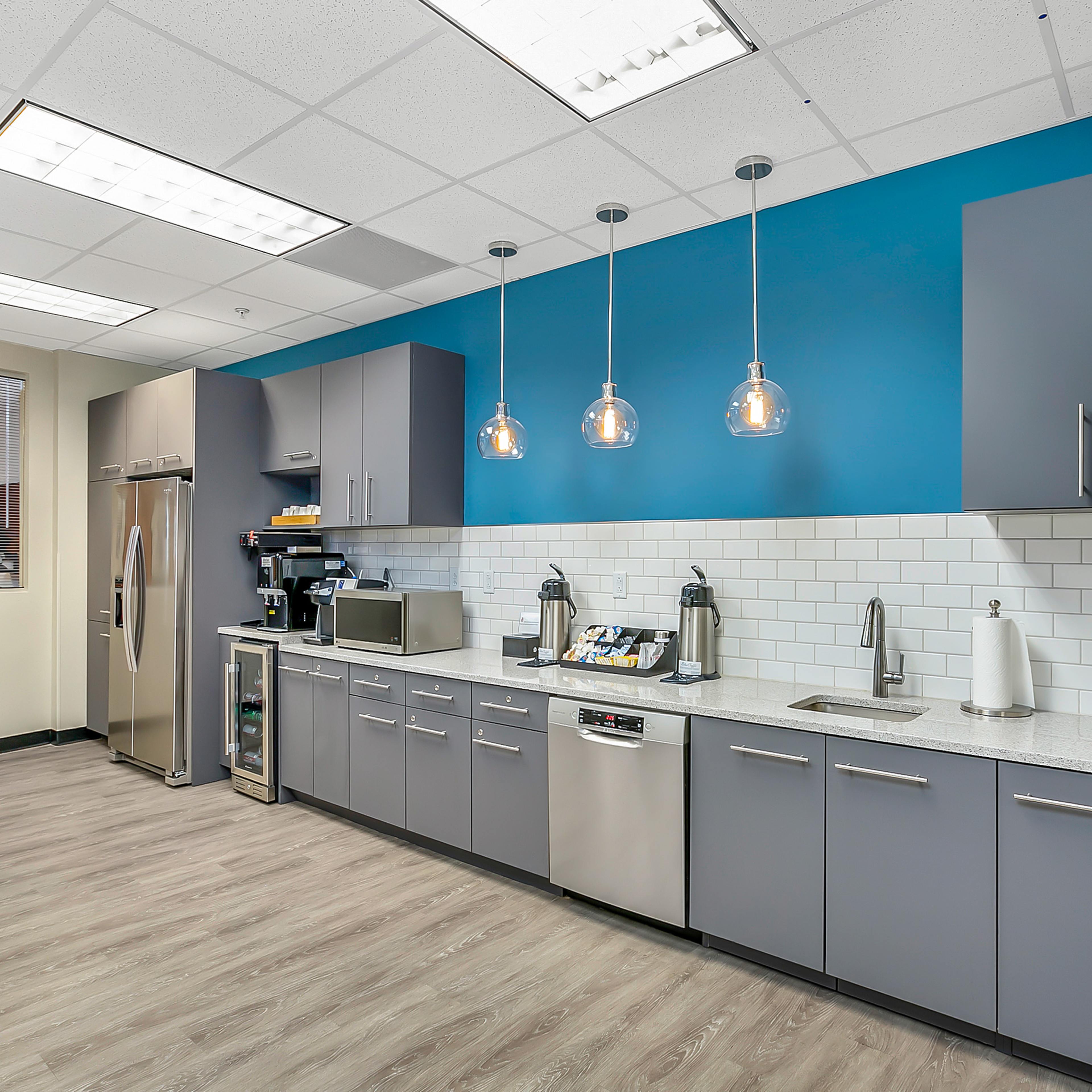 A modern kitchen with gray cabinets, a stainless steel refrigerator, and pendant lighting over a countertop with a coffee maker and various kitchen appliances.