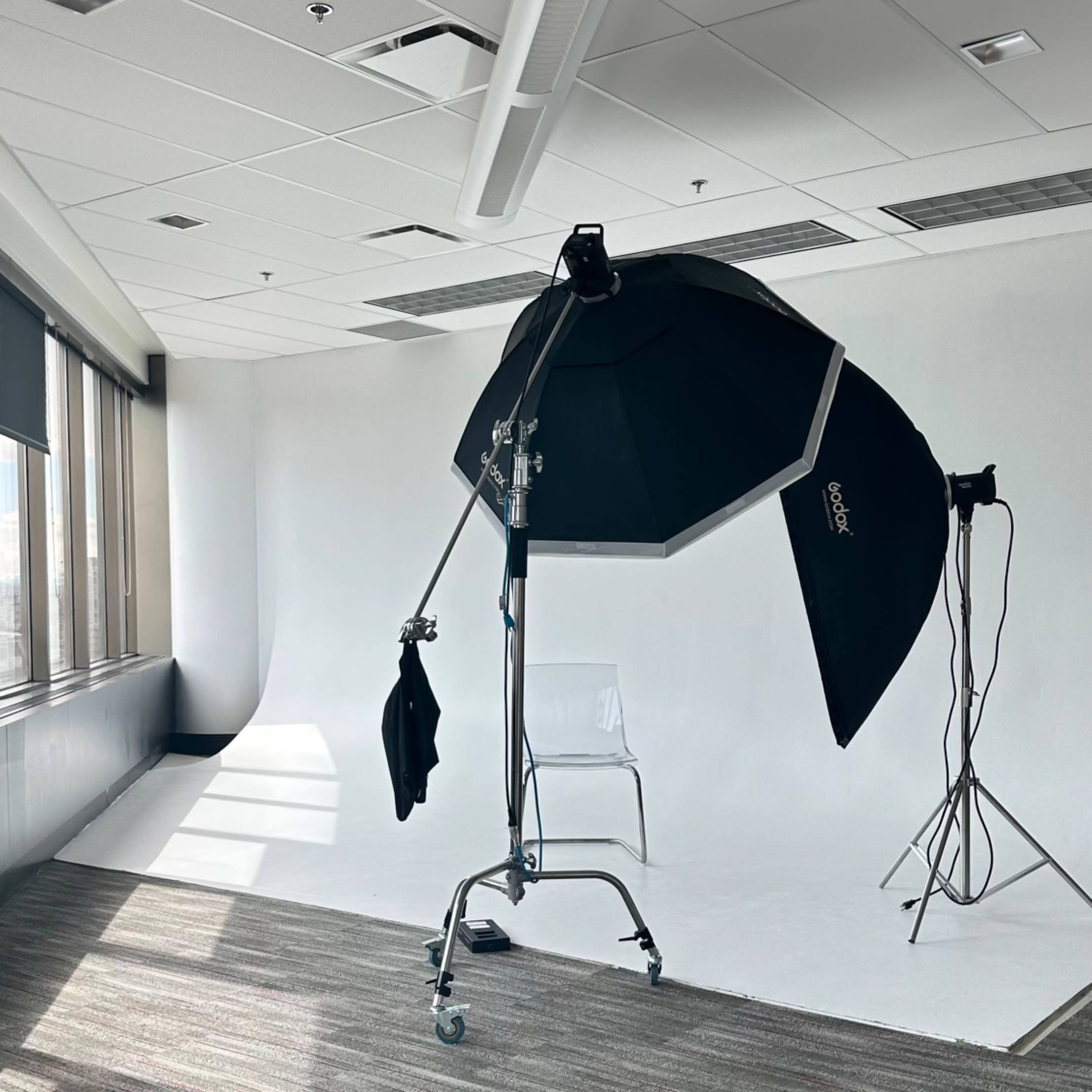 A photography studio setup features a large softbox on a stand and a backdrop against a white wall with a patterned carpet.