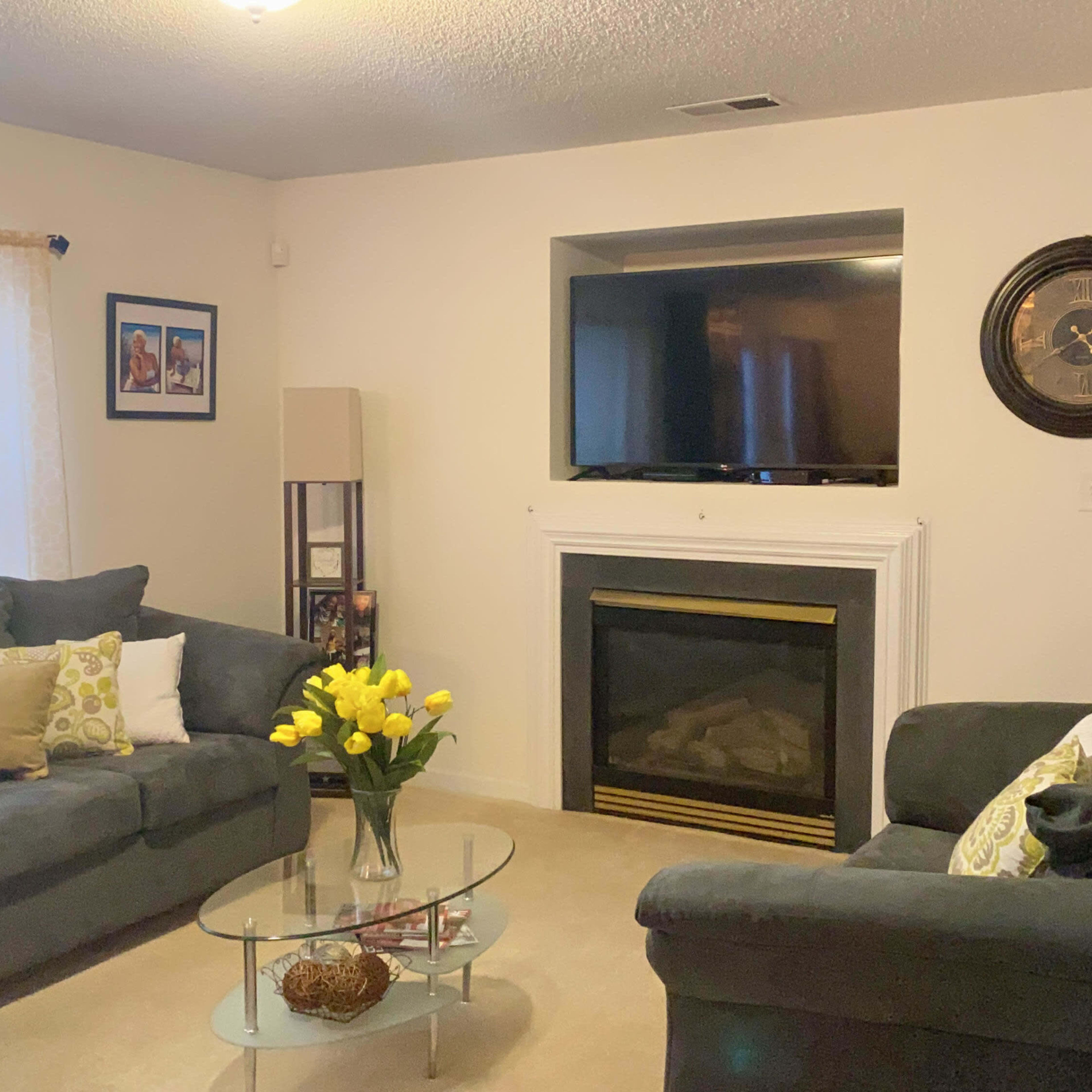 The living room features two gray sofas facing a glass coffee table, with a fireplace and a television mounted above it.