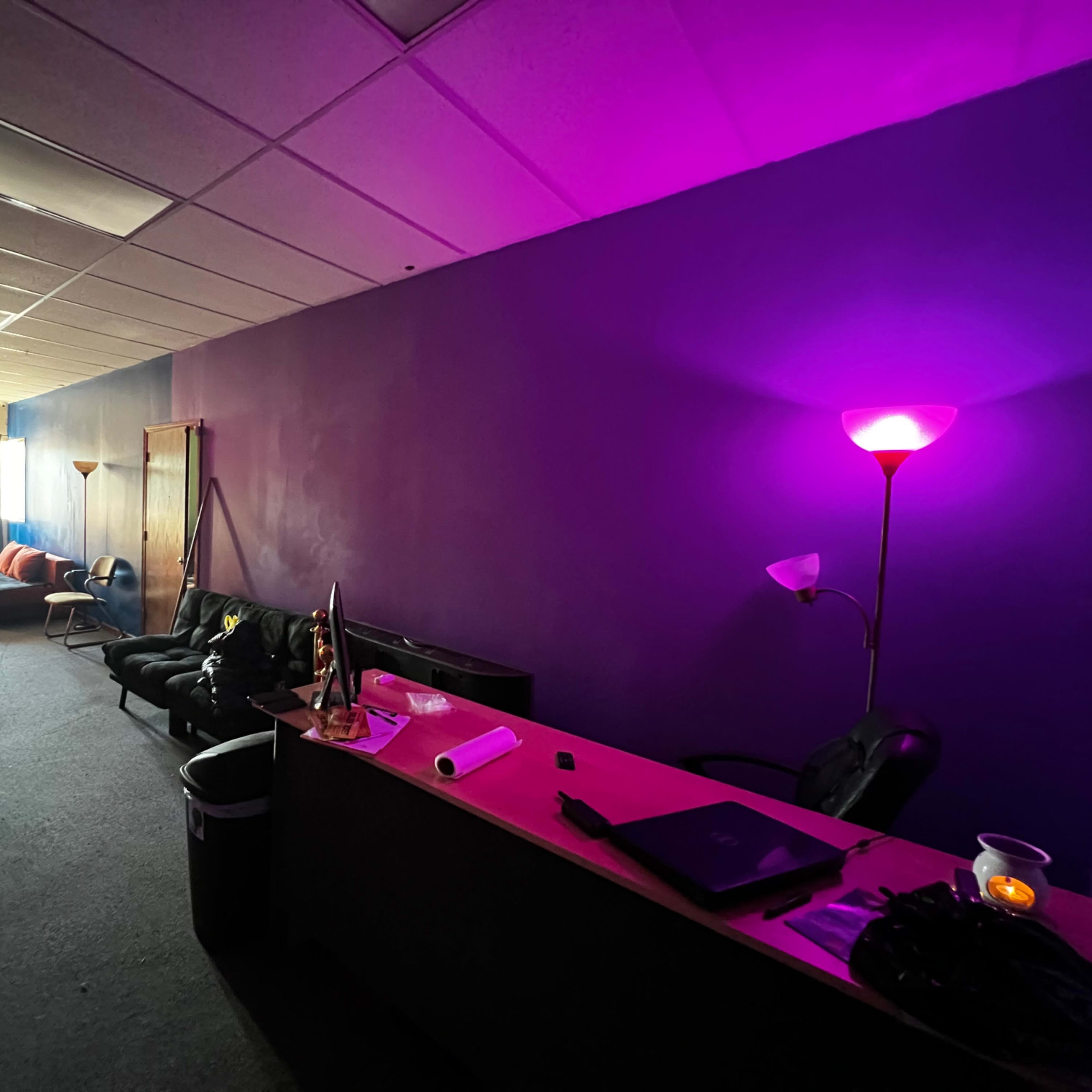 A brightly colored room with a dark purple wall, a black couch, a desk with items on it, and a lamp providing light on the right side.