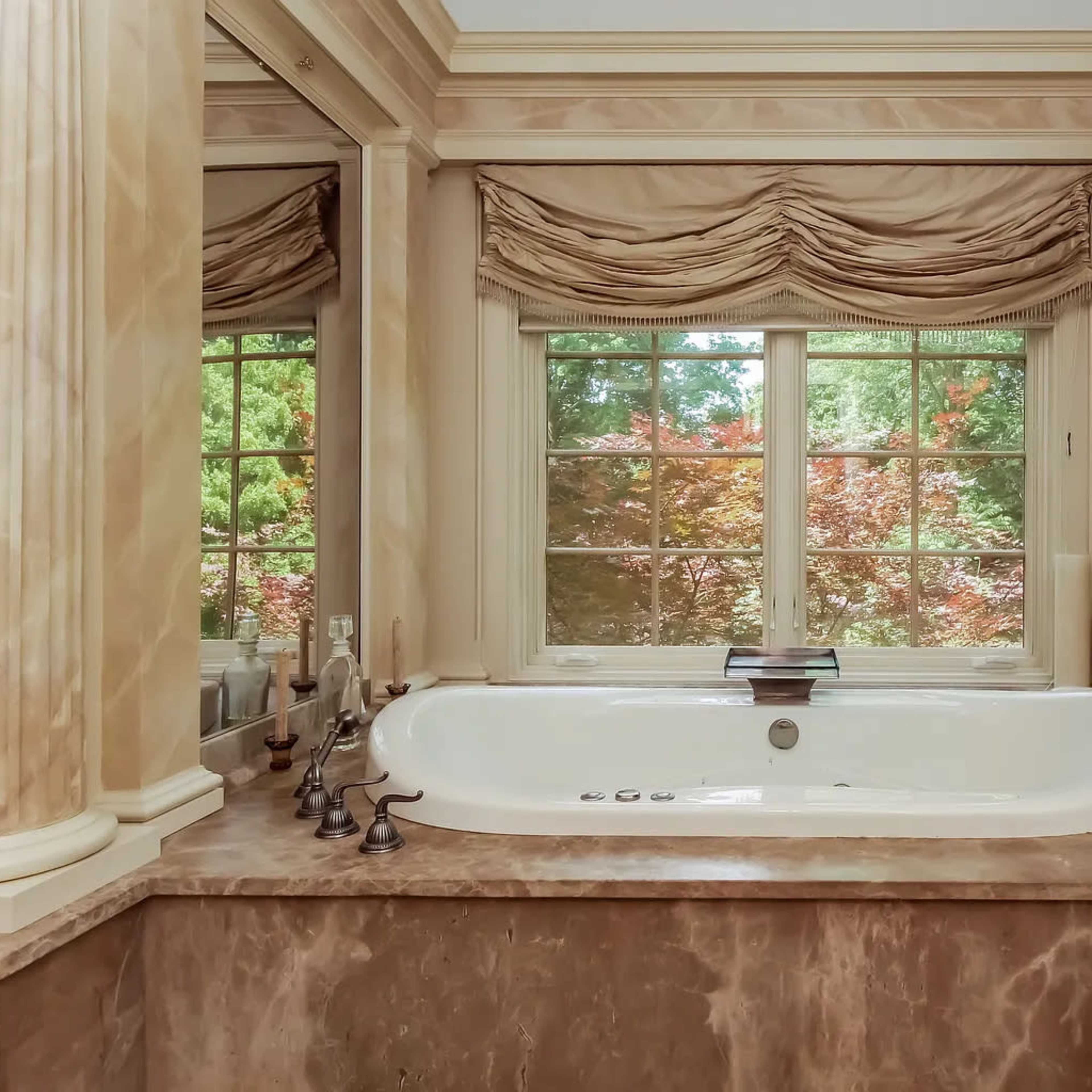 A spacious bathroom features a white tub surrounded by marble finishes and large windows offering a view of lush greenery outside.