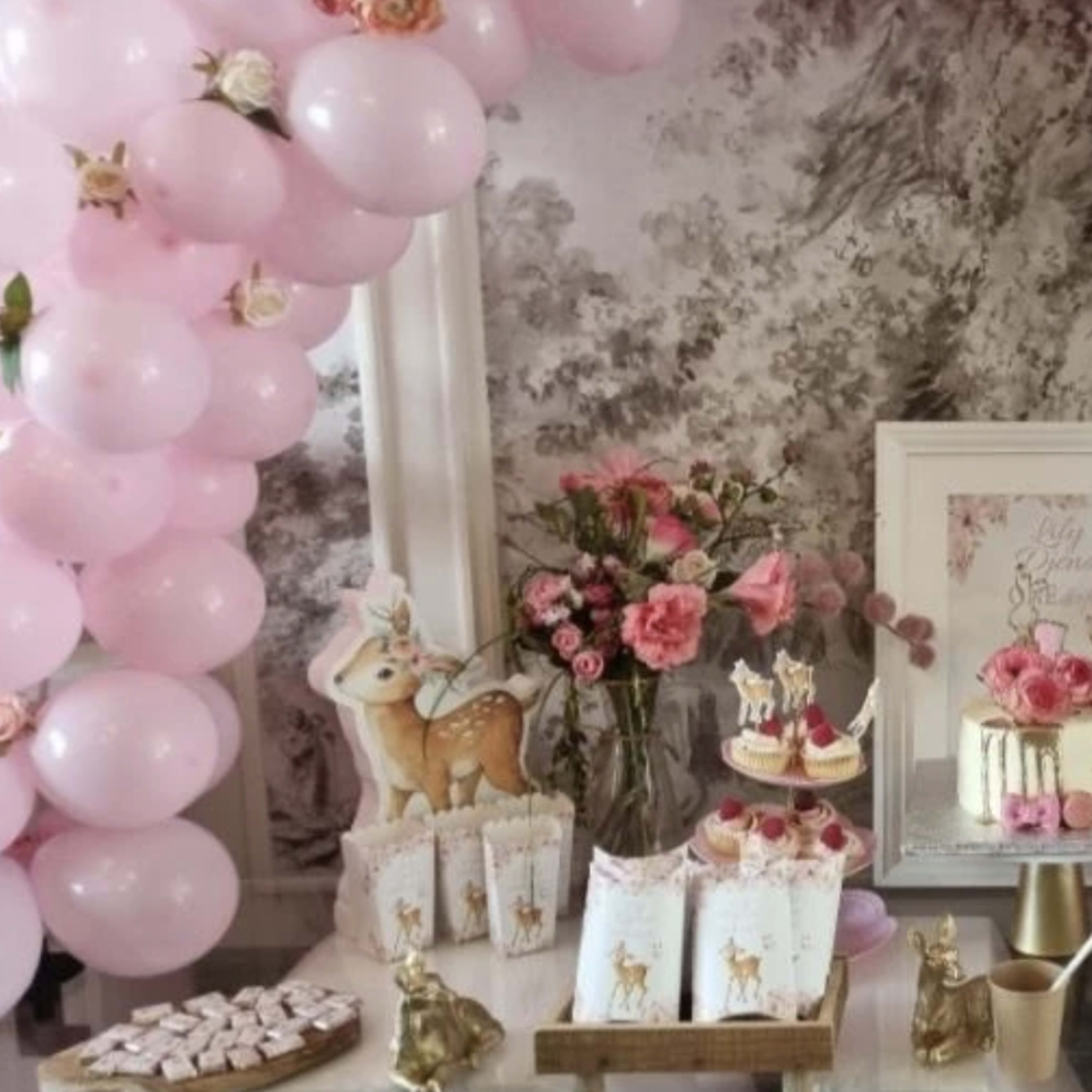 A dessert table is decorated with pink balloons, a floral arrangement, a cake, and various sweets, all set against a vintage wallpaper backdrop.