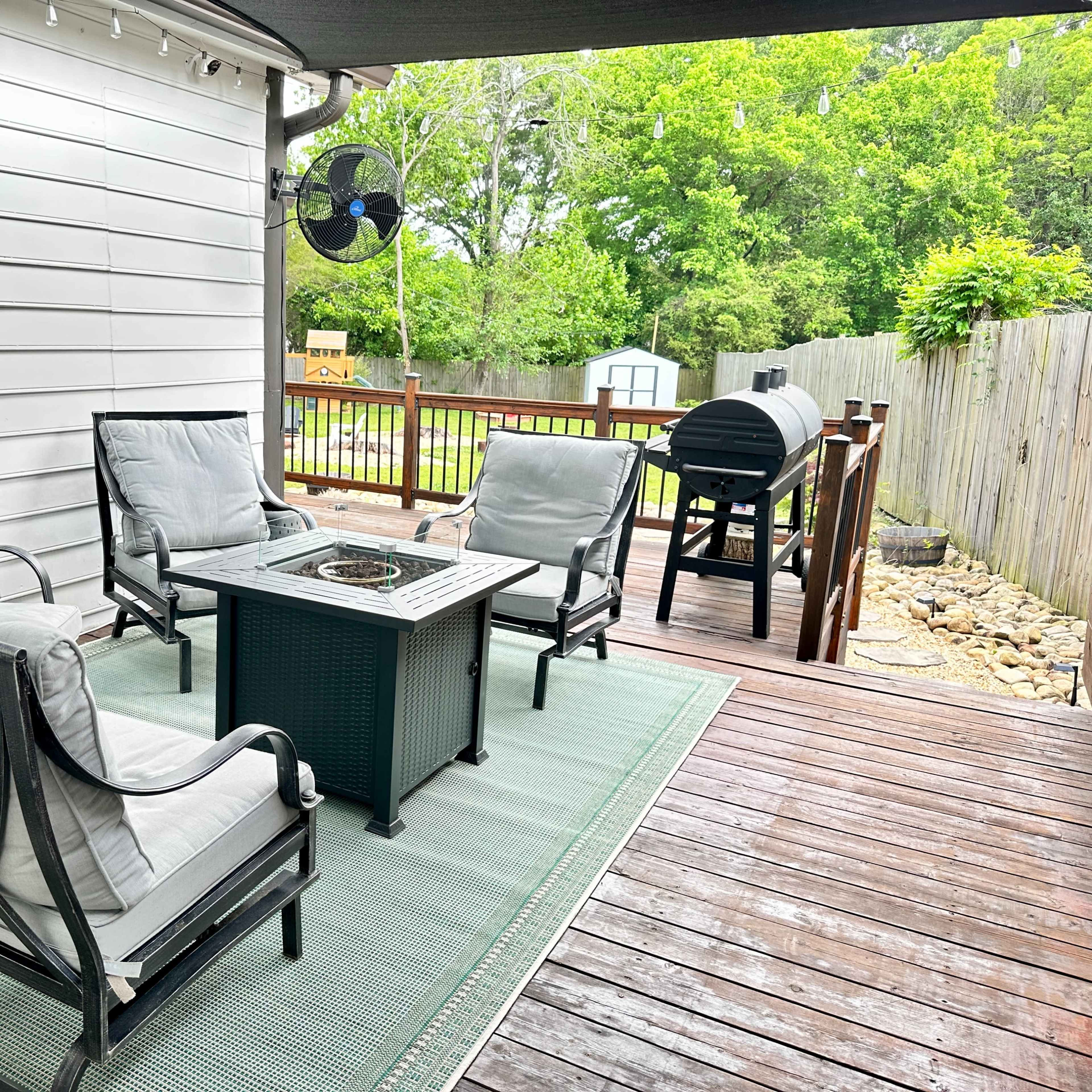 A patio area features four chairs arranged around a fire pit table on a wooden deck, with a grill and greenery visible in the background.