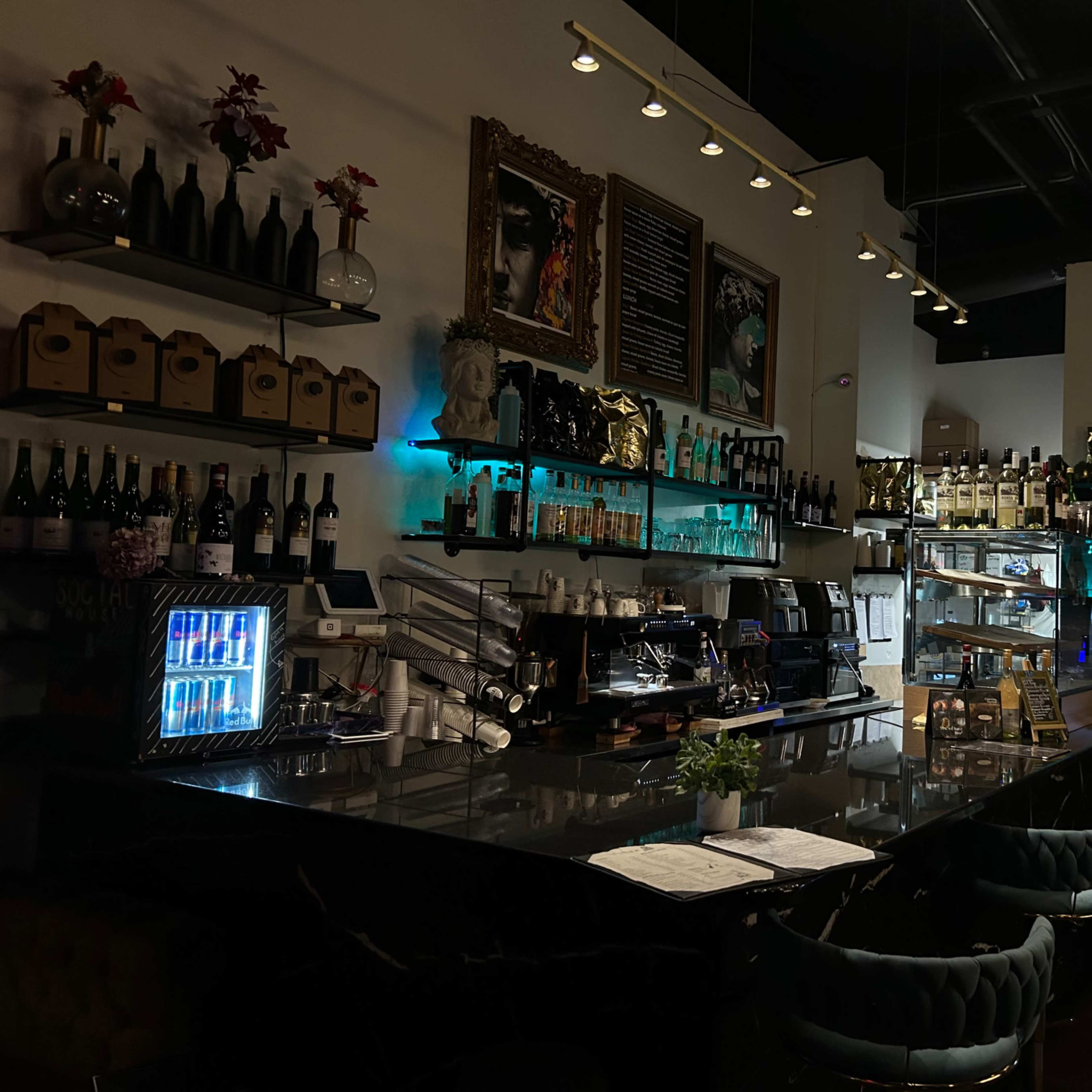 A dimly lit bar with shelves of bottles, a coffee machine, and a counter displaying menus and a chalkboard sign.