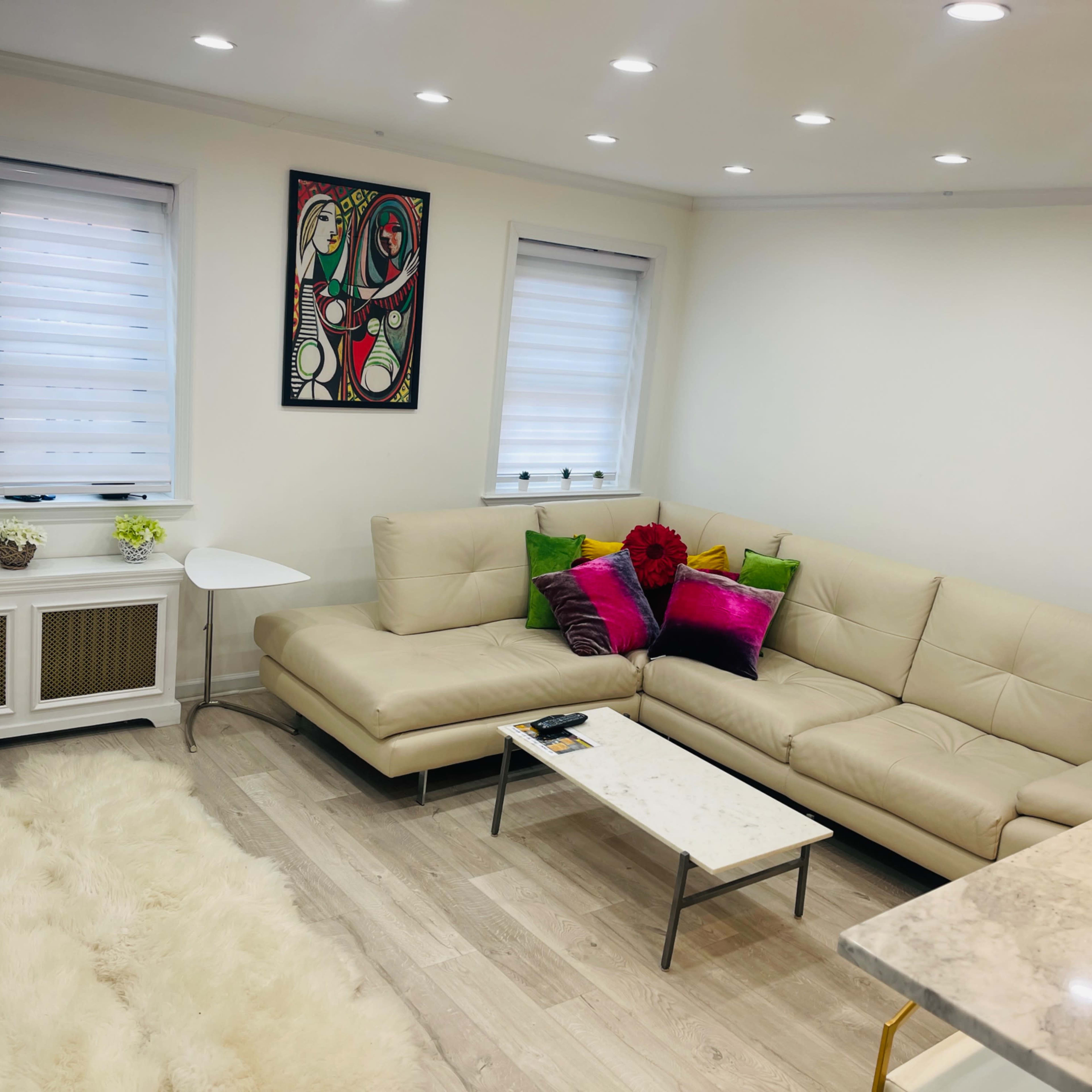 A modern living room featuring a beige leather sectional sofa with colorful cushions, a small white side table, and a marble-topped coffee table atop a light-colored wooden floor.