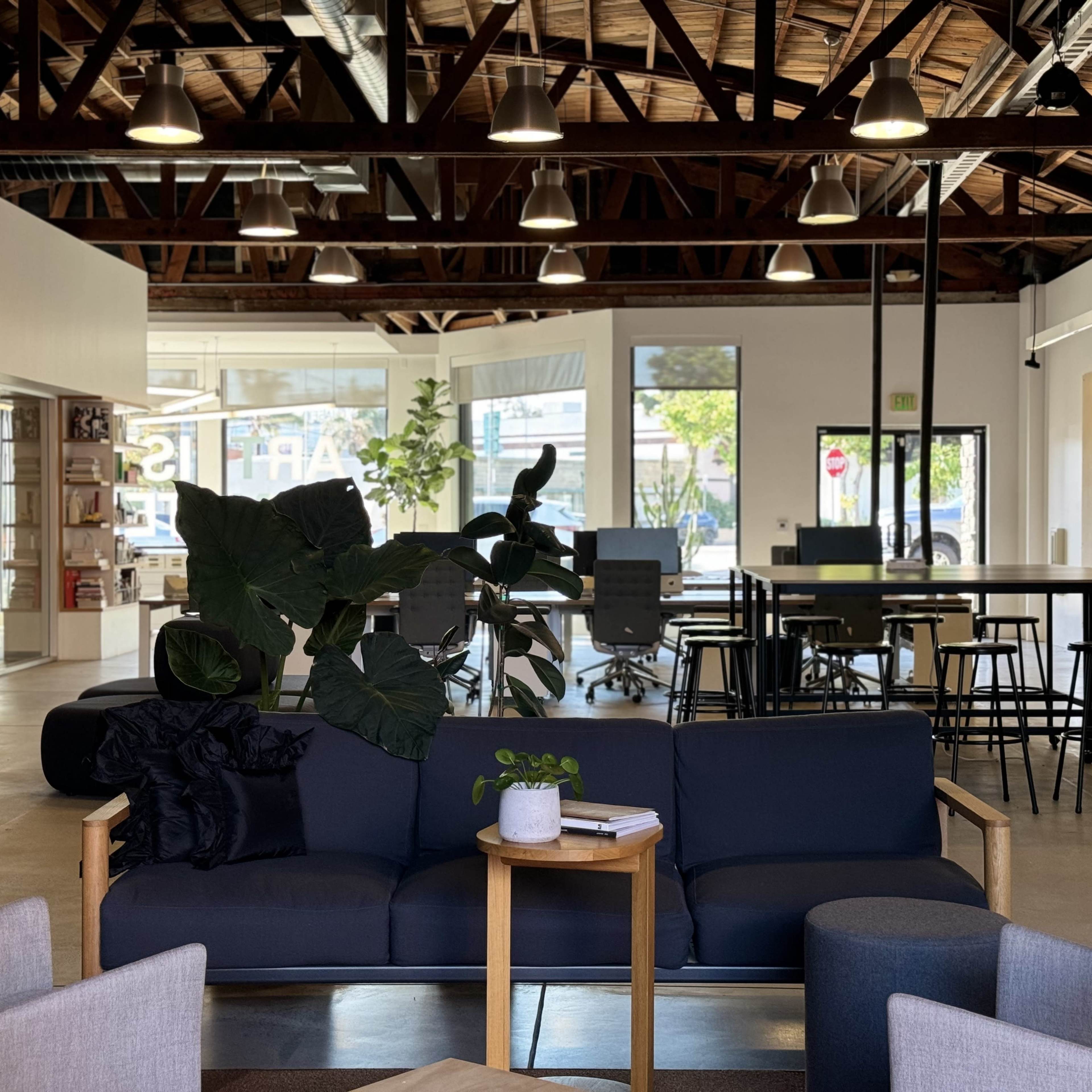 A modern office space featuring a seating area with a dark sofa and small table, surrounded by workstations and large windows.