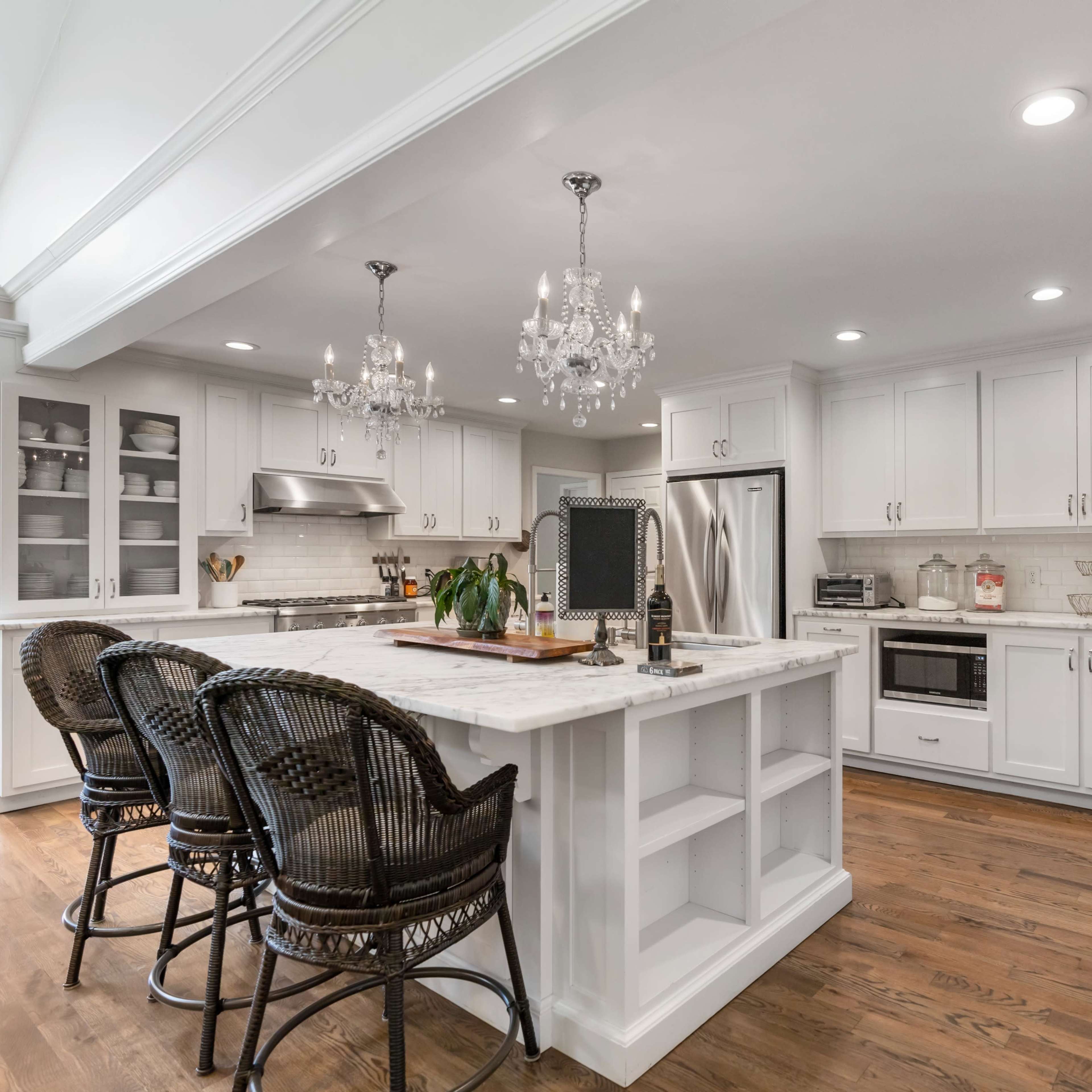A modern kitchen features white cabinetry, a large marble island with seating, and stainless steel appliances, illuminated by decorative chandeliers.