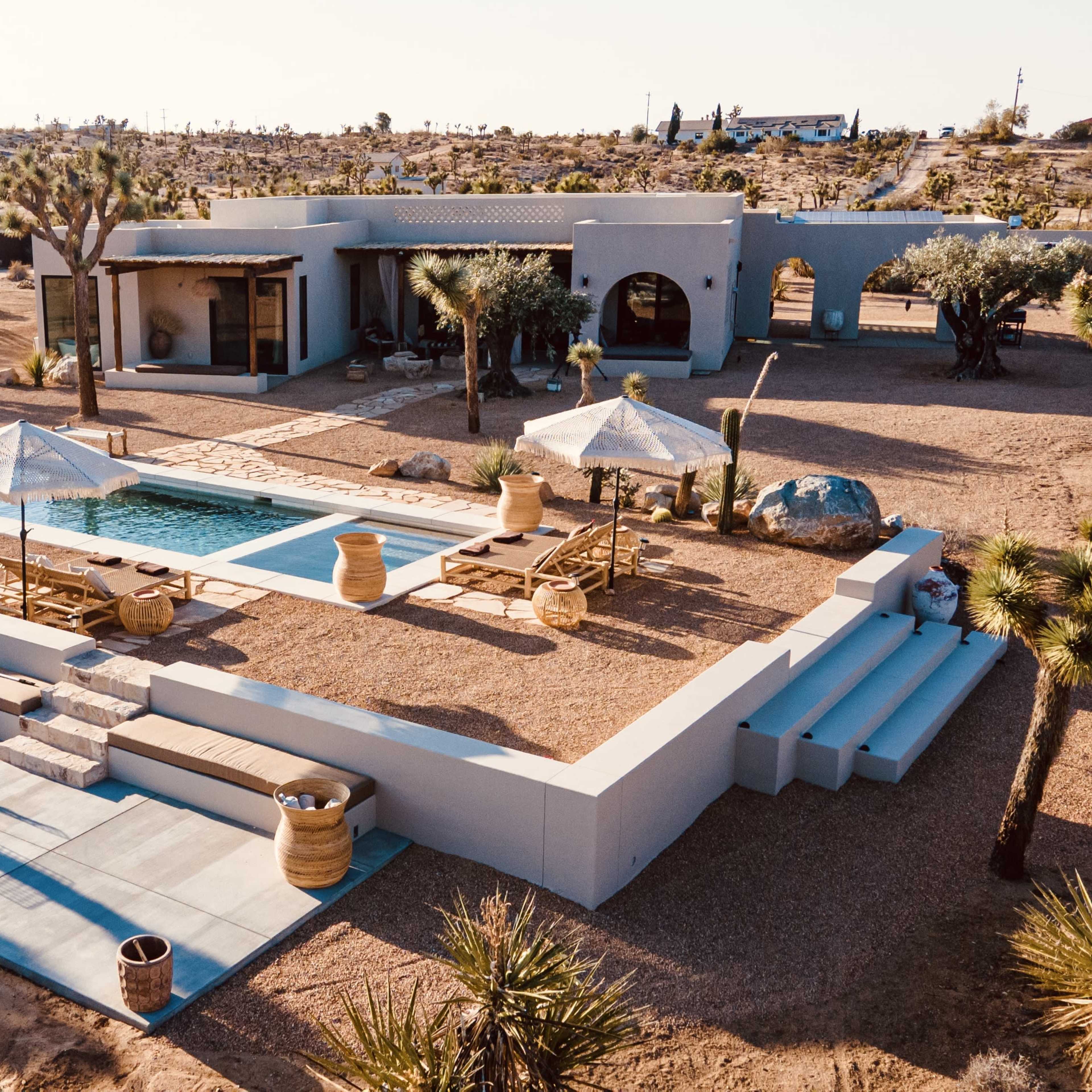 A modern desert house with a pool, surrounded by landscaping featuring Joshua trees and outdoor seating areas.