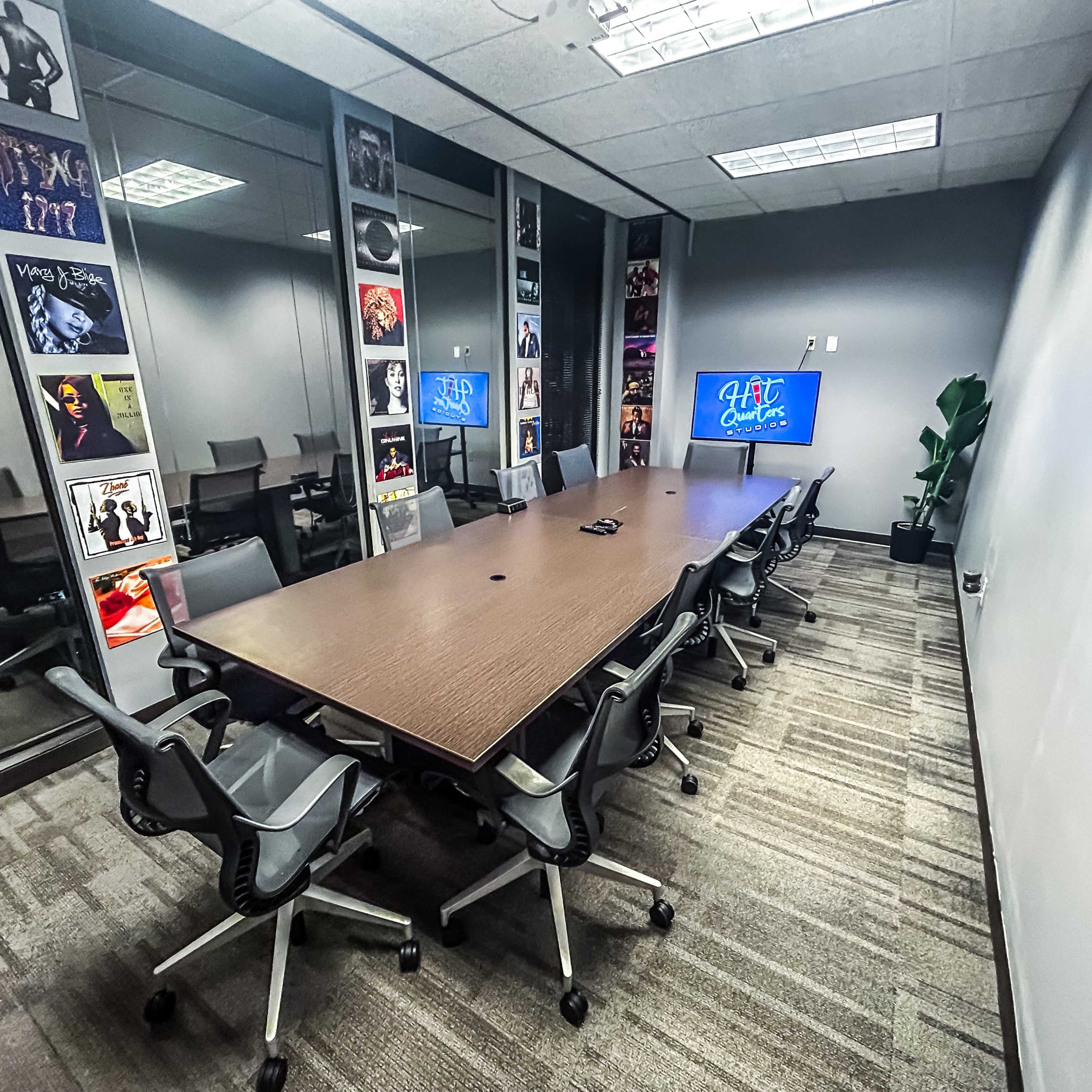 A modern conference room features a large wooden table surrounded by ergonomic chairs, with glass walls displaying framed posters and a television screen.