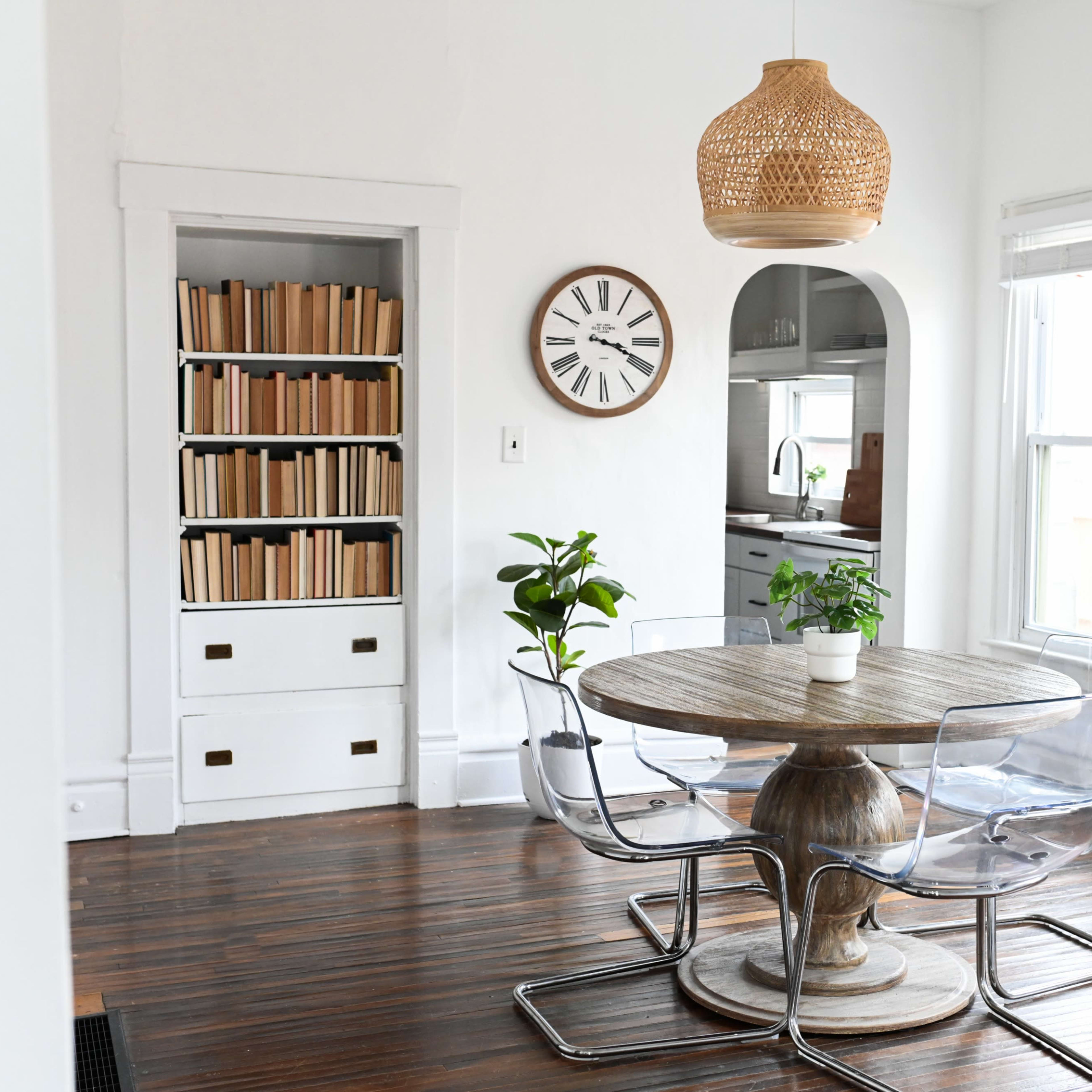 A bright dining area with a round wooden table surrounded by transparent chairs, a clock on the wall, and a bookshelf filled with books.