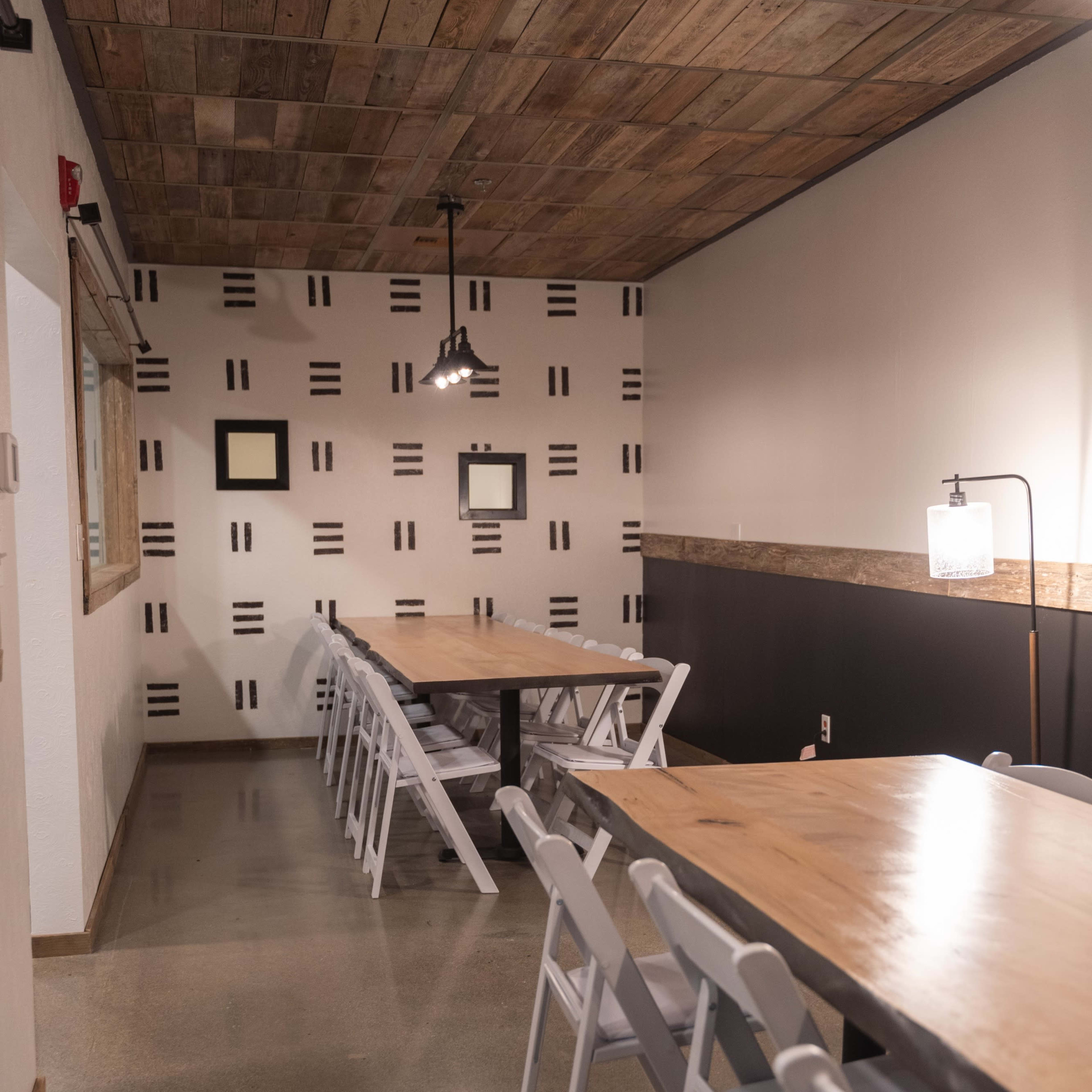 A long room features two tables with wooden tops and white folding chairs arranged neatly, along with a patterned accent wall and a rustic ceiling.