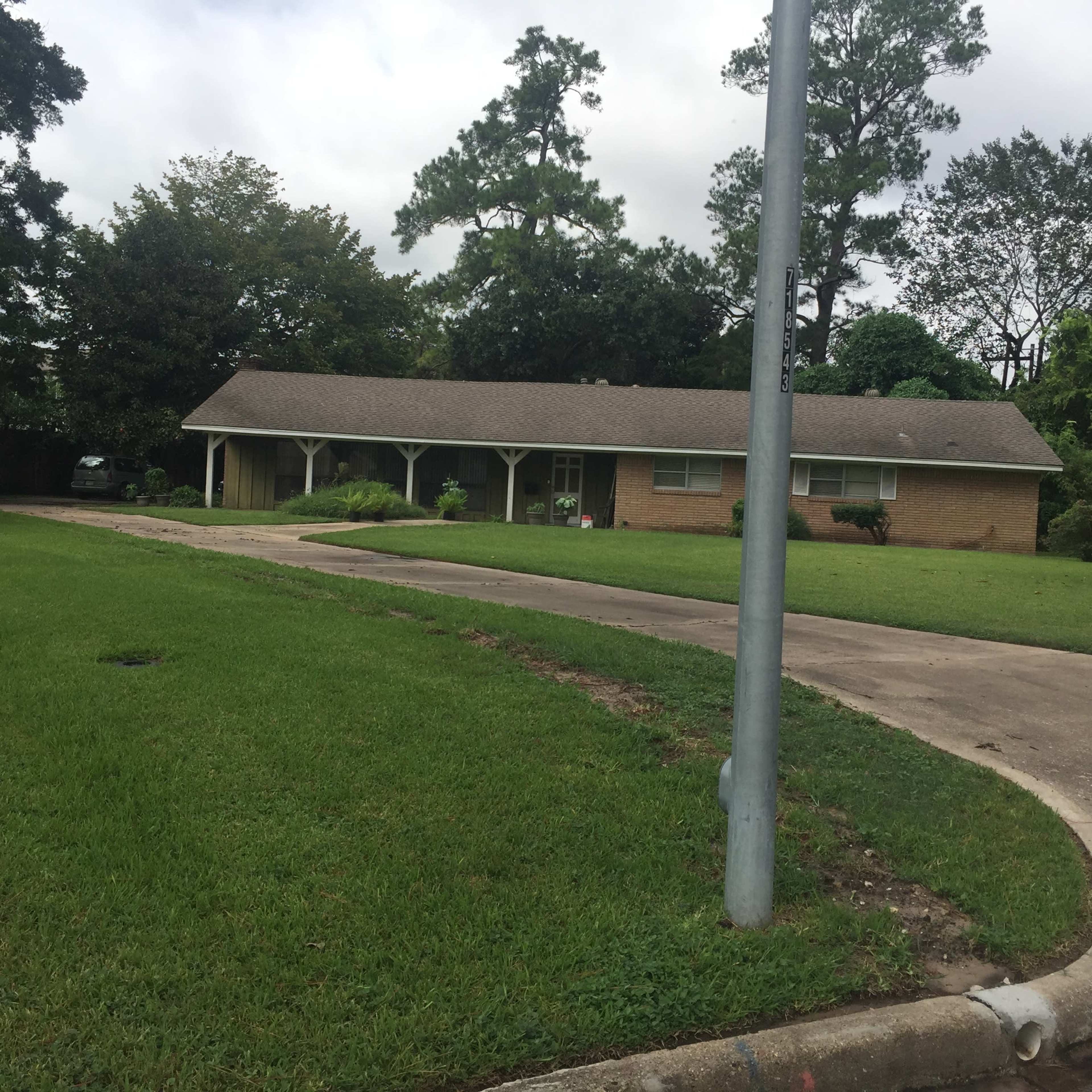 A single-story brick house with a front porch set on a grassy lawn, bordered by trees and a paved driveway.