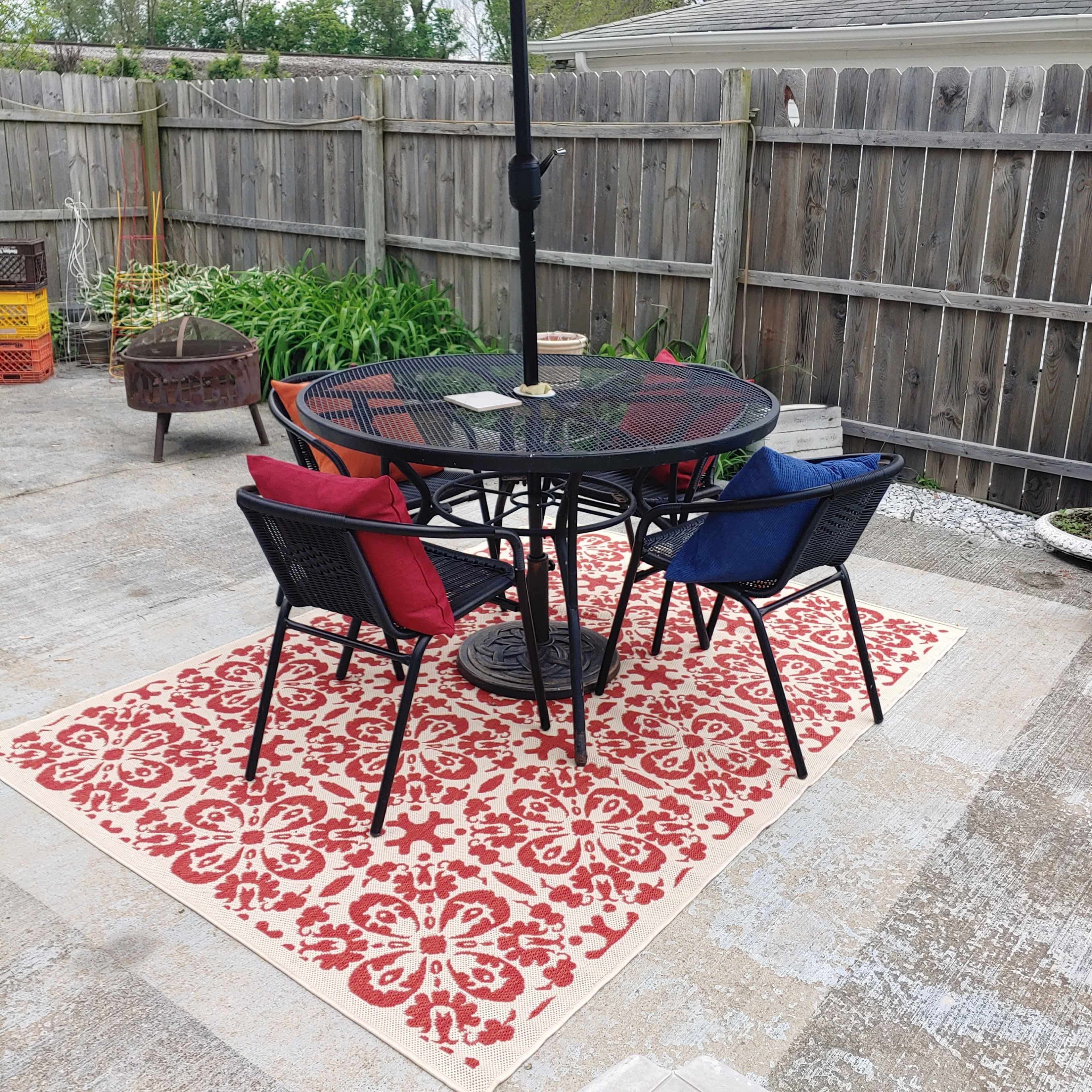 A round table with two chairs is set on a patterned rug beneath a red umbrella in a fenced outdoor area.