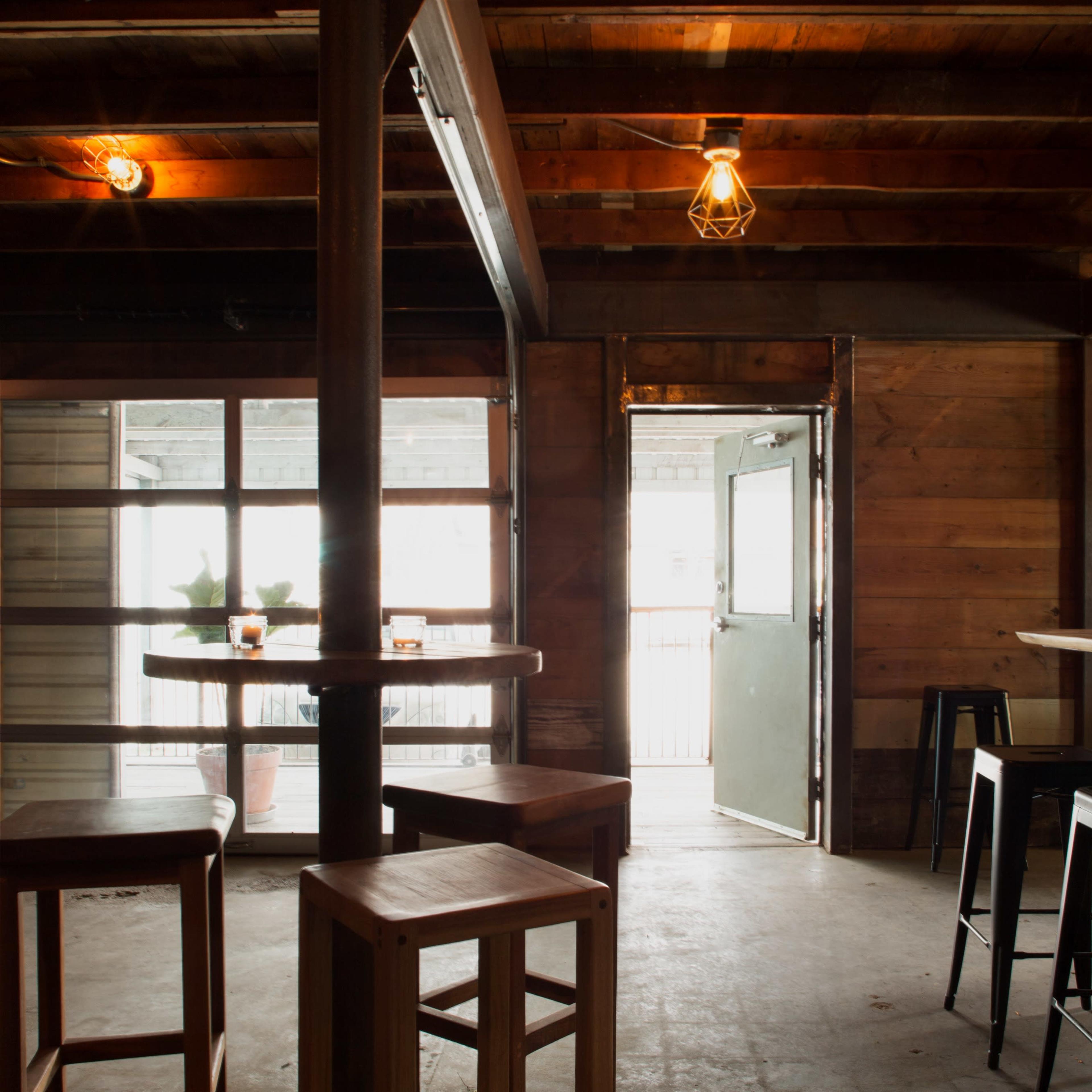 A rustic bar interior featuring wooden beams, metal stools, and an open door leading outside.
