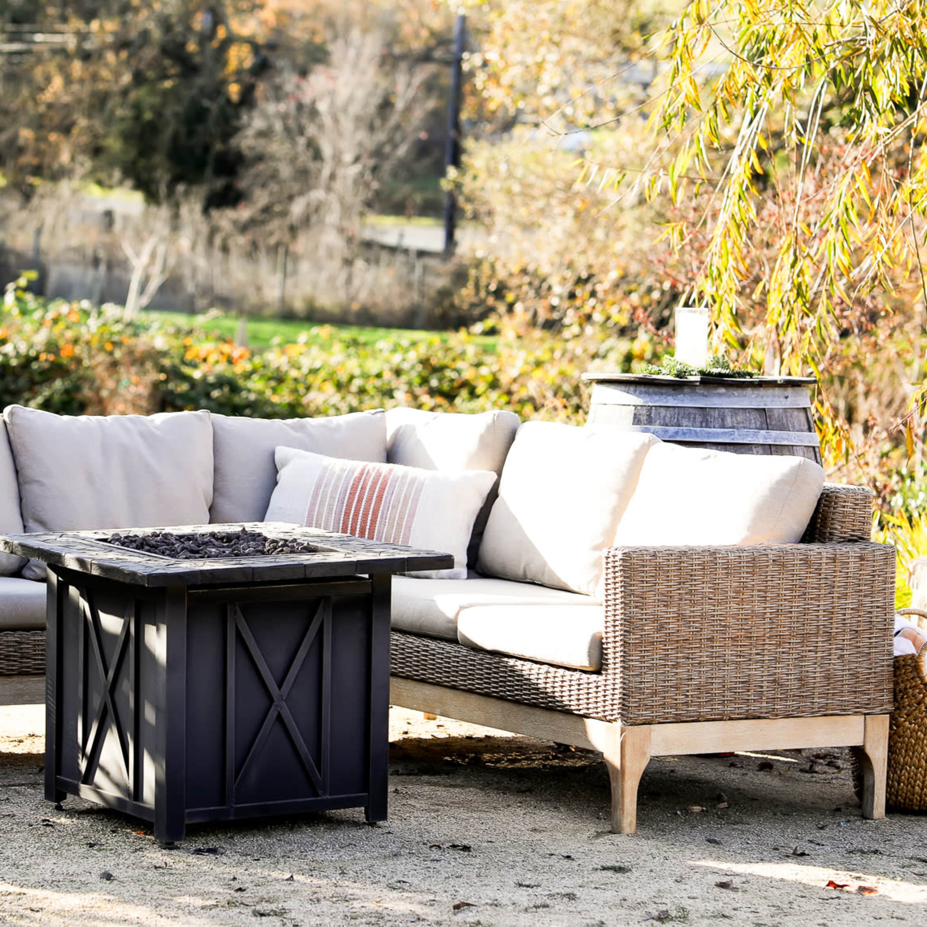 A patio seating area features a wicker sectional sofa and a black fire pit table surrounded by greenery.