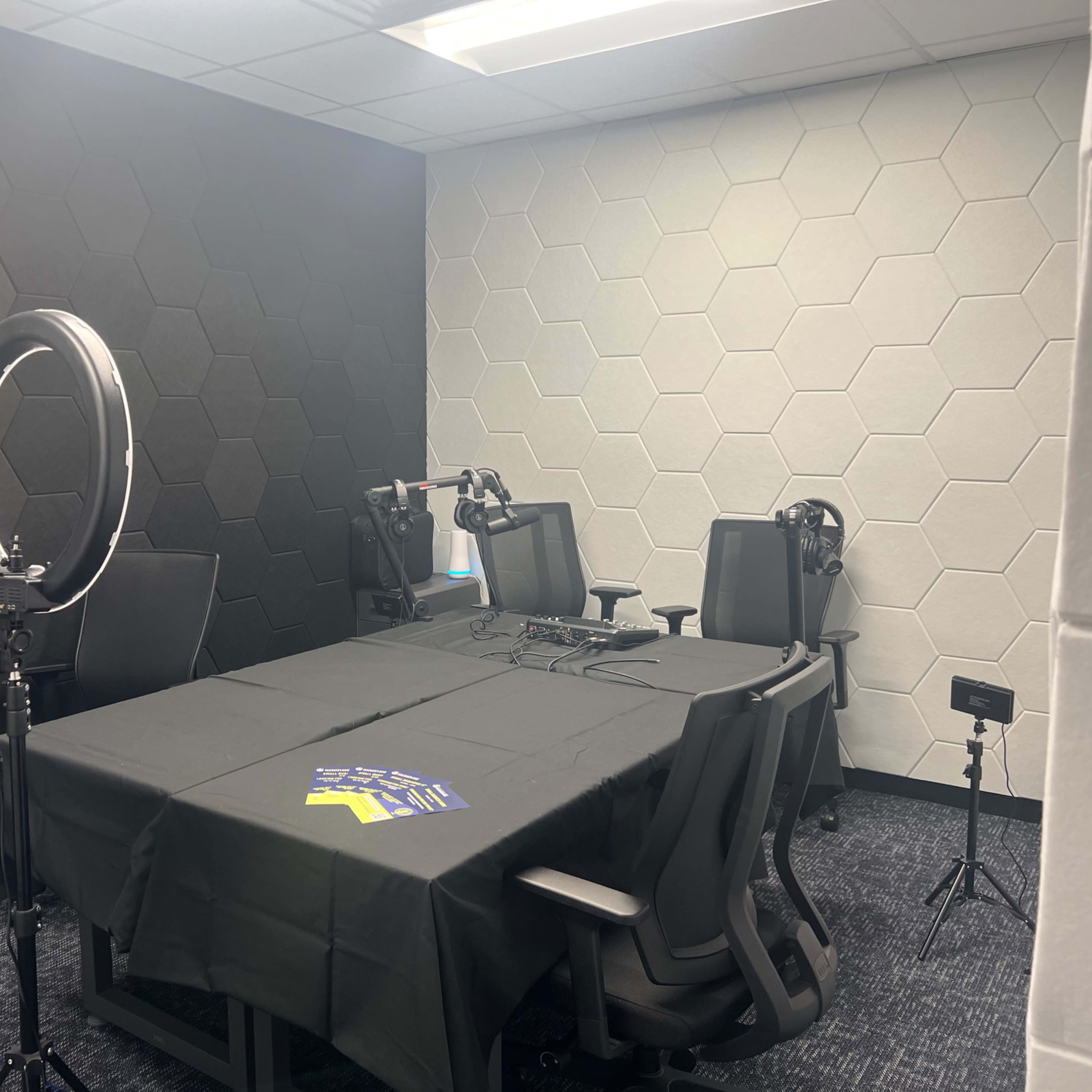 A meeting room with hexagonal patterned walls, a large table covered with a black tablecloth, and equipment such as microphones and lights set up for recording.