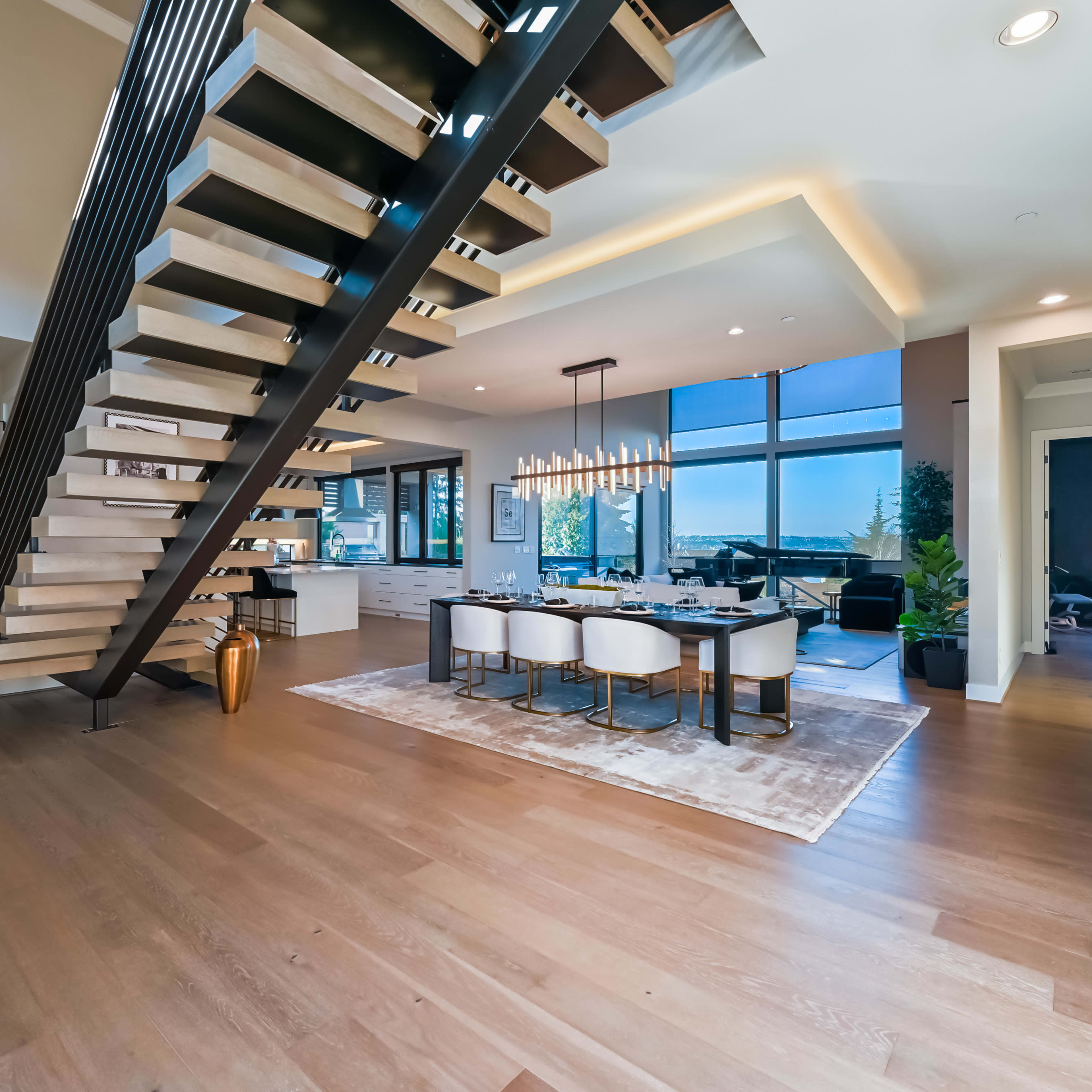 A modern interior space with an open floor plan featuring a staircase, a dining area with a table and chairs, and large windows offering a view.