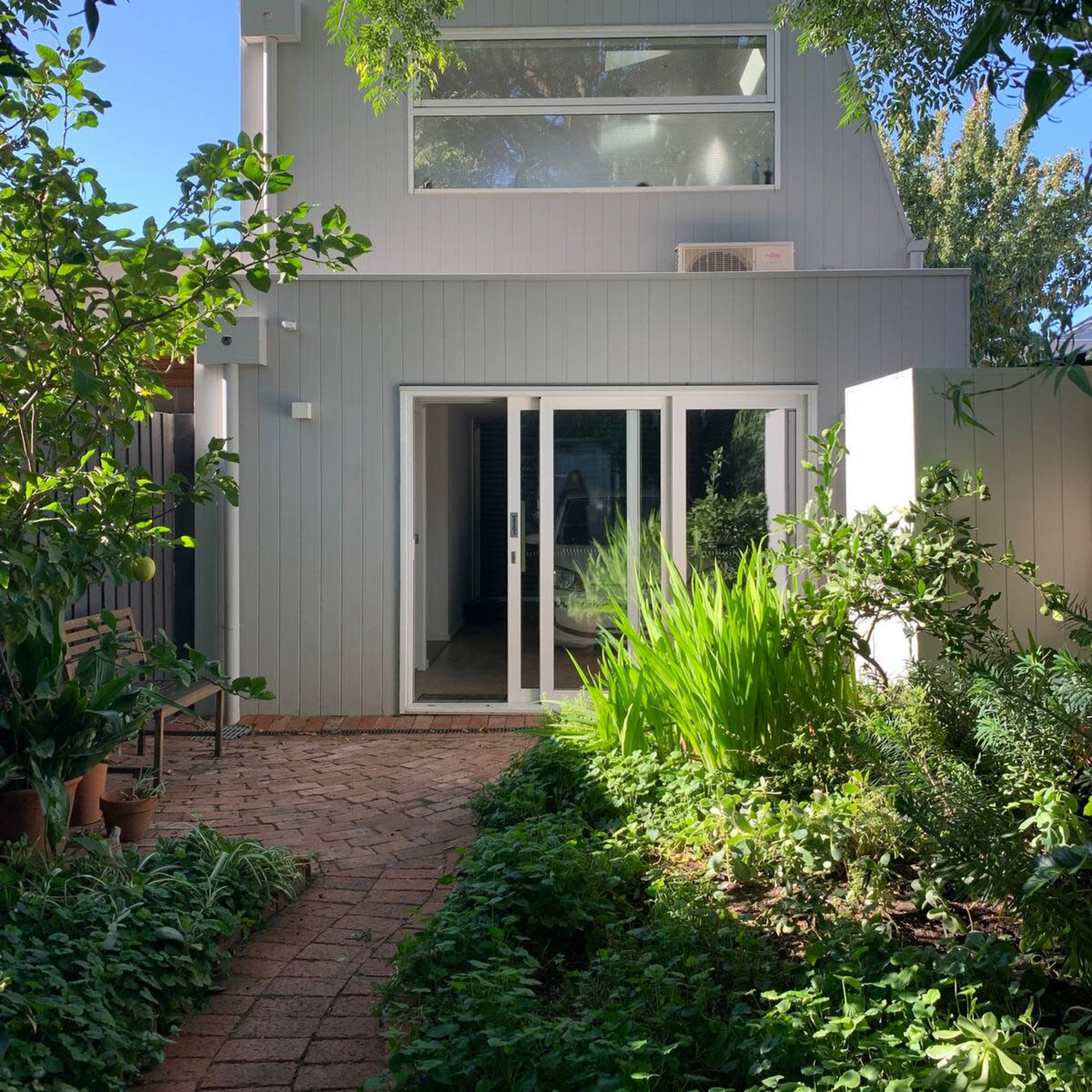 A light gray house with large windows, surrounded by lush greenery and a pathway made of bricks.
