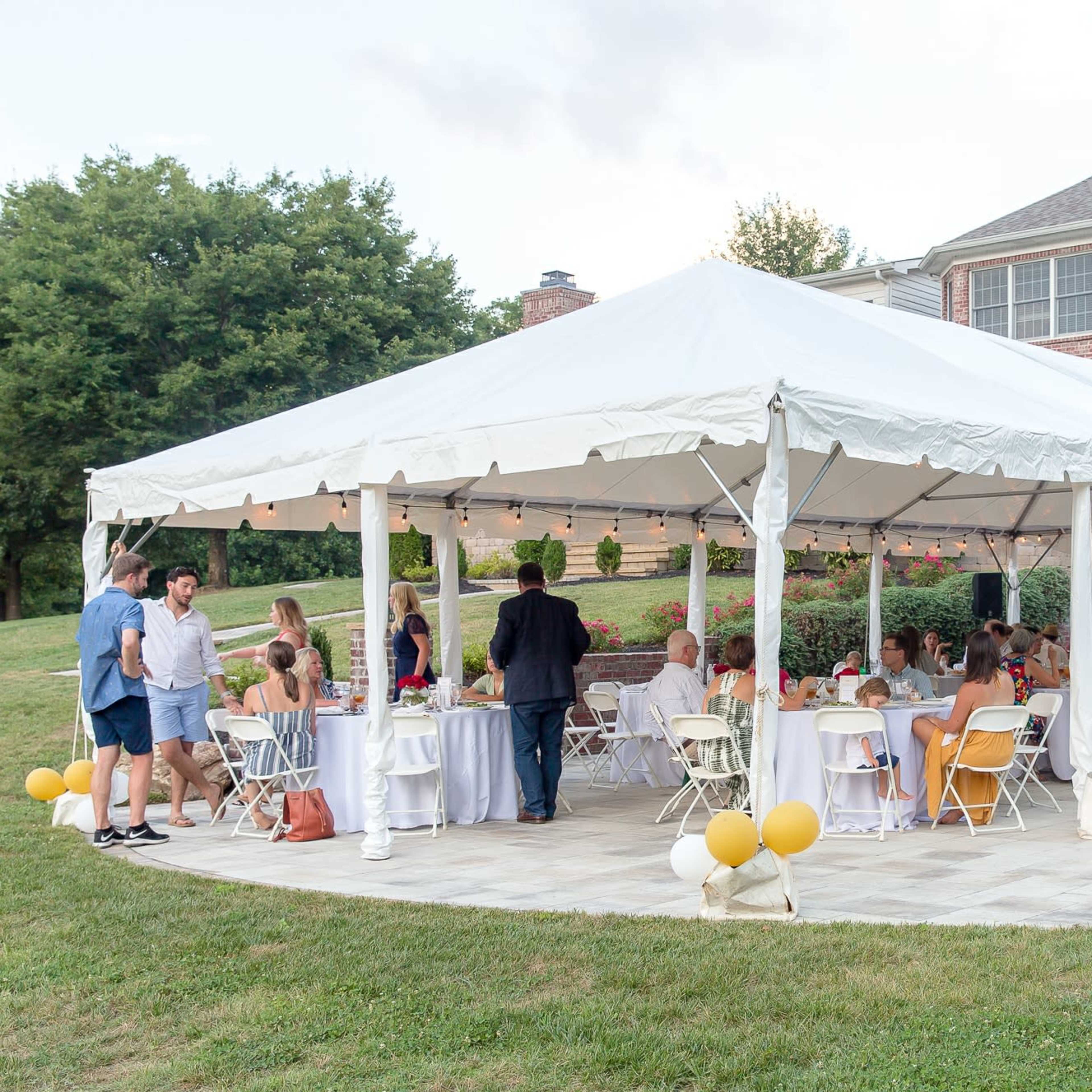 A large outdoor tent filled with guests seated at tables is set up beside a brick house for a gathering.