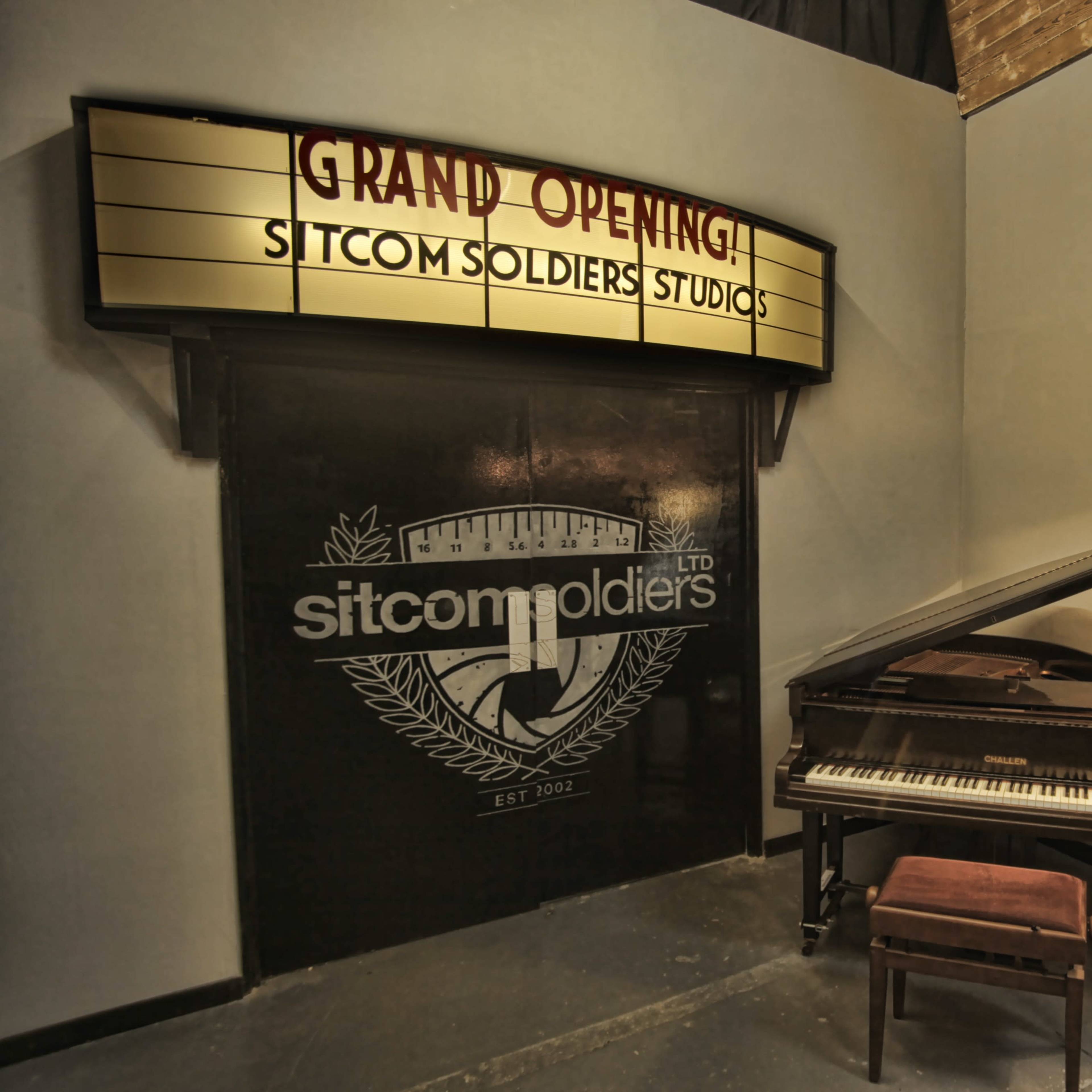 A corner of a studio adorned with a sign reading "GRAND OPENING!" above a grand piano and a brown chair.