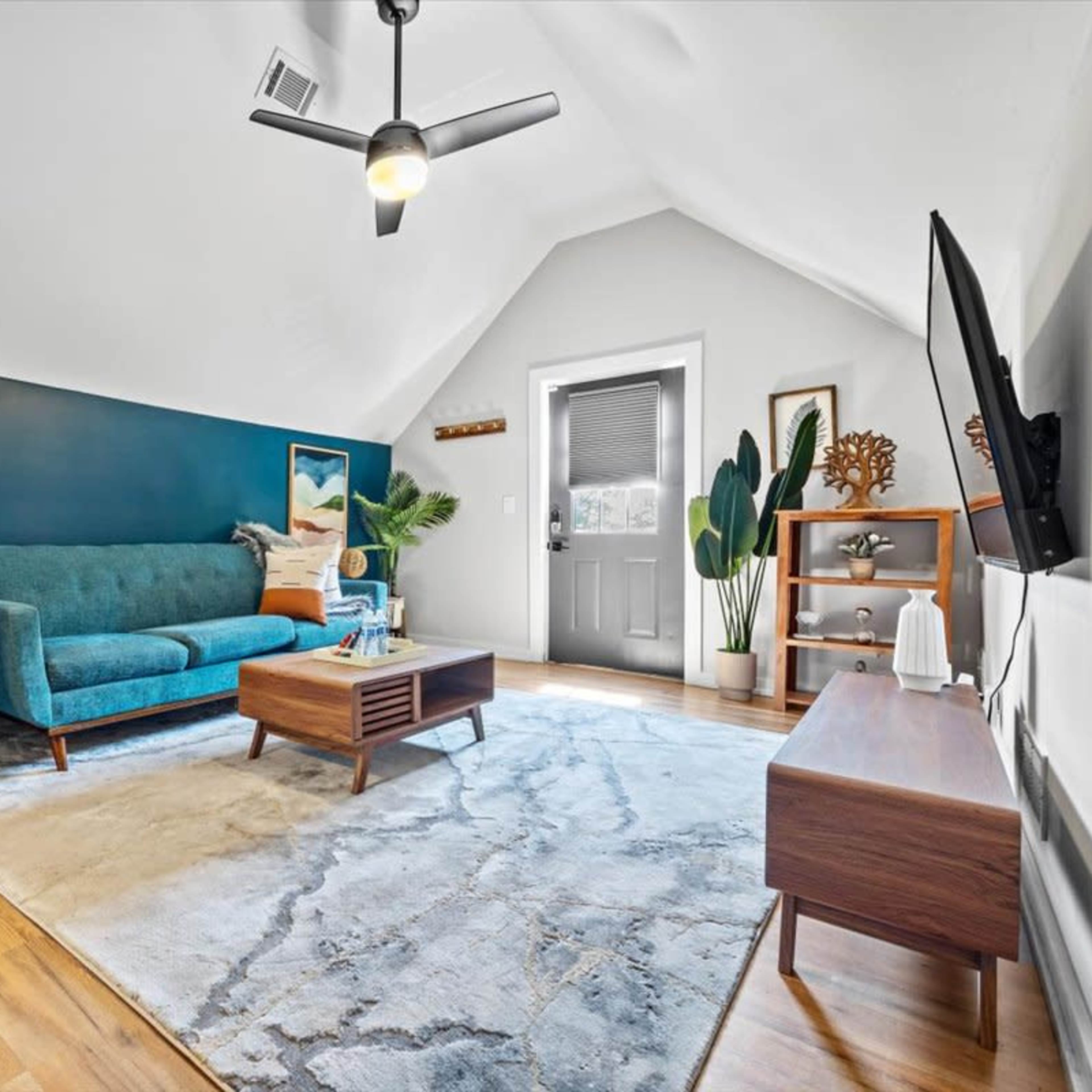 The room features a teal sofa and a wooden coffee table on a large area rug, with a door leading outside and wall art hanging above the furniture.