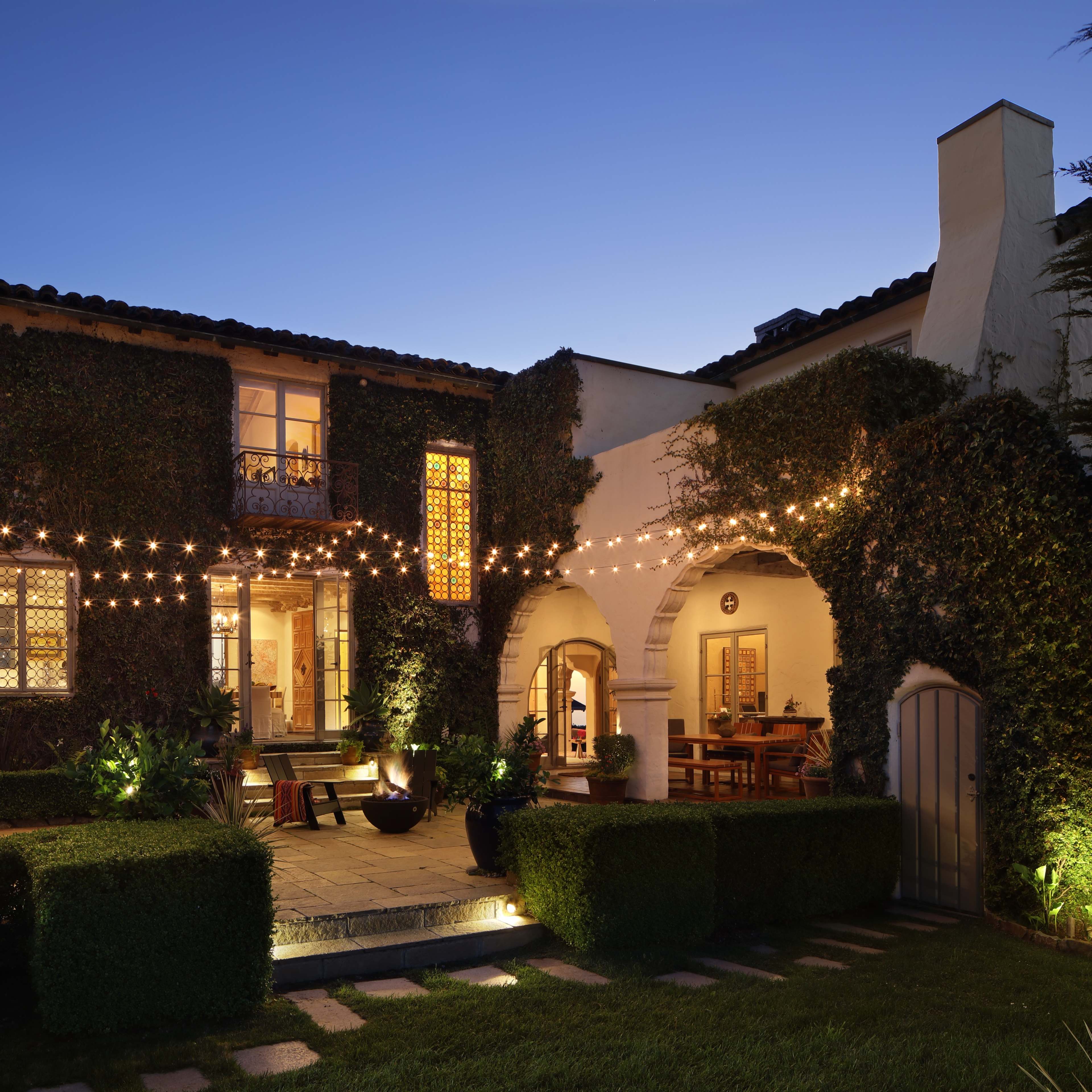A two-story house with ivy-covered walls is illuminated by string lights in the backyard at dusk.