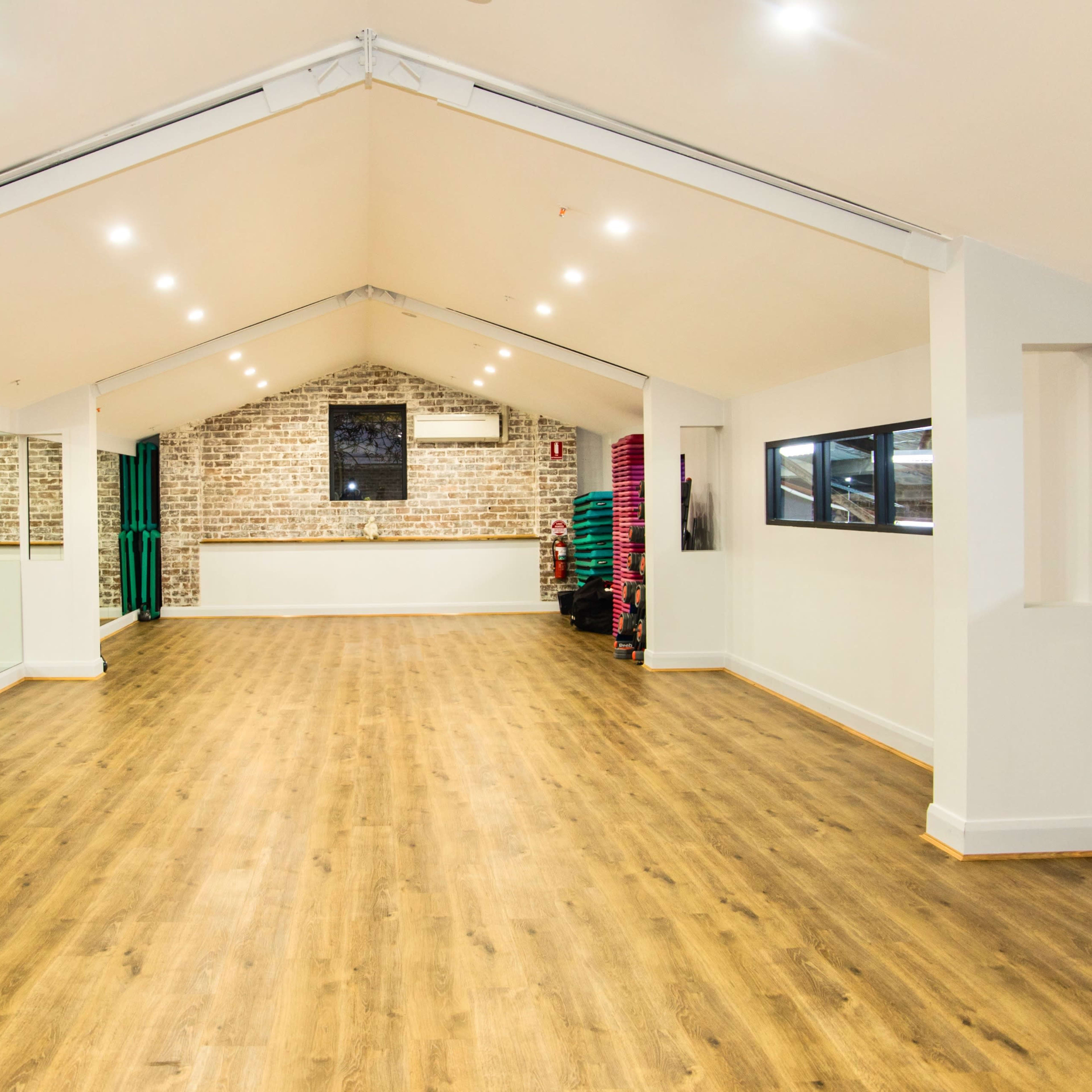 A spacious fitness studio with wooden floors, white walls, and ample natural light from large windows.