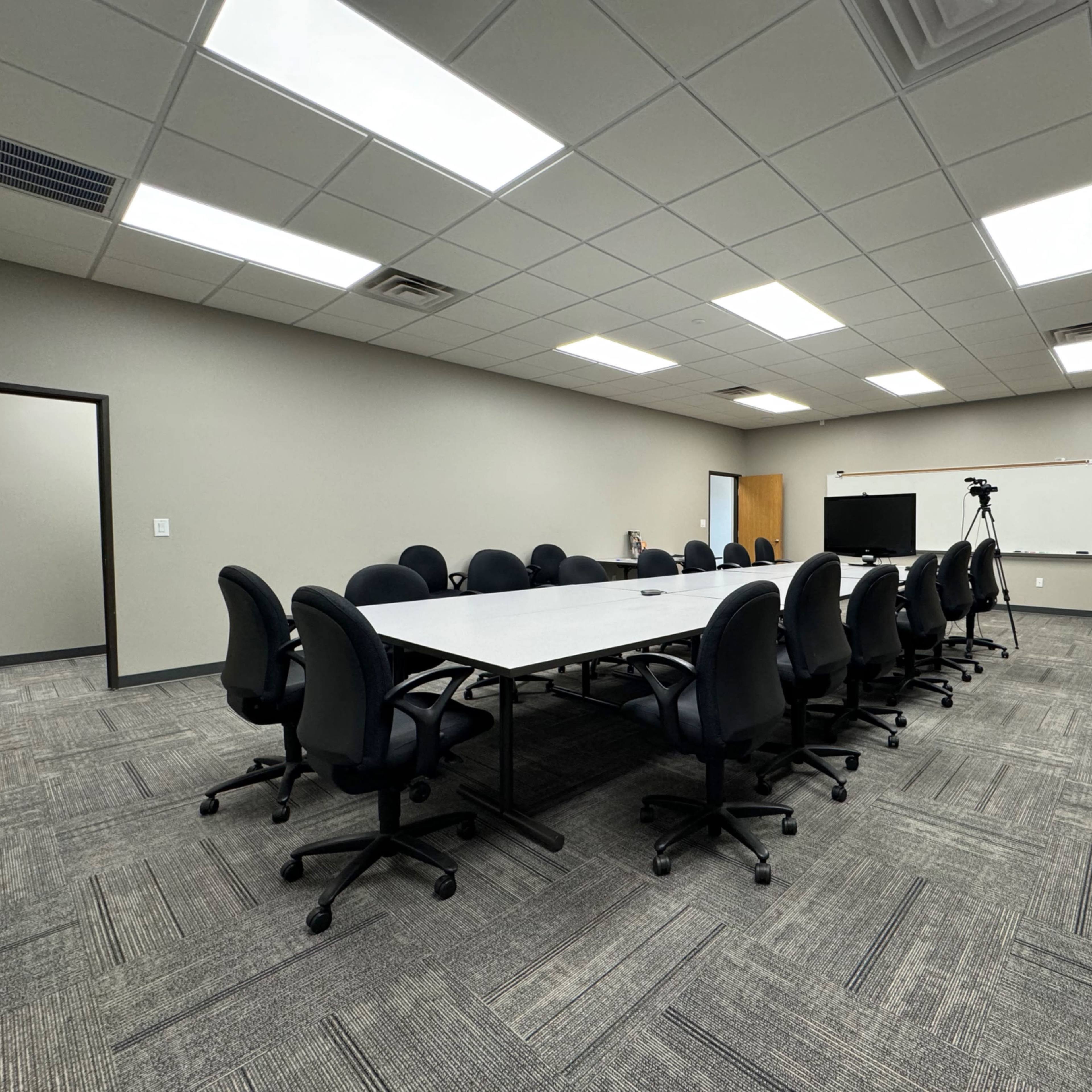 A spacious conference room features a long rectangular table surrounded by black rolling chairs, with additional chairs aligned against the walls.