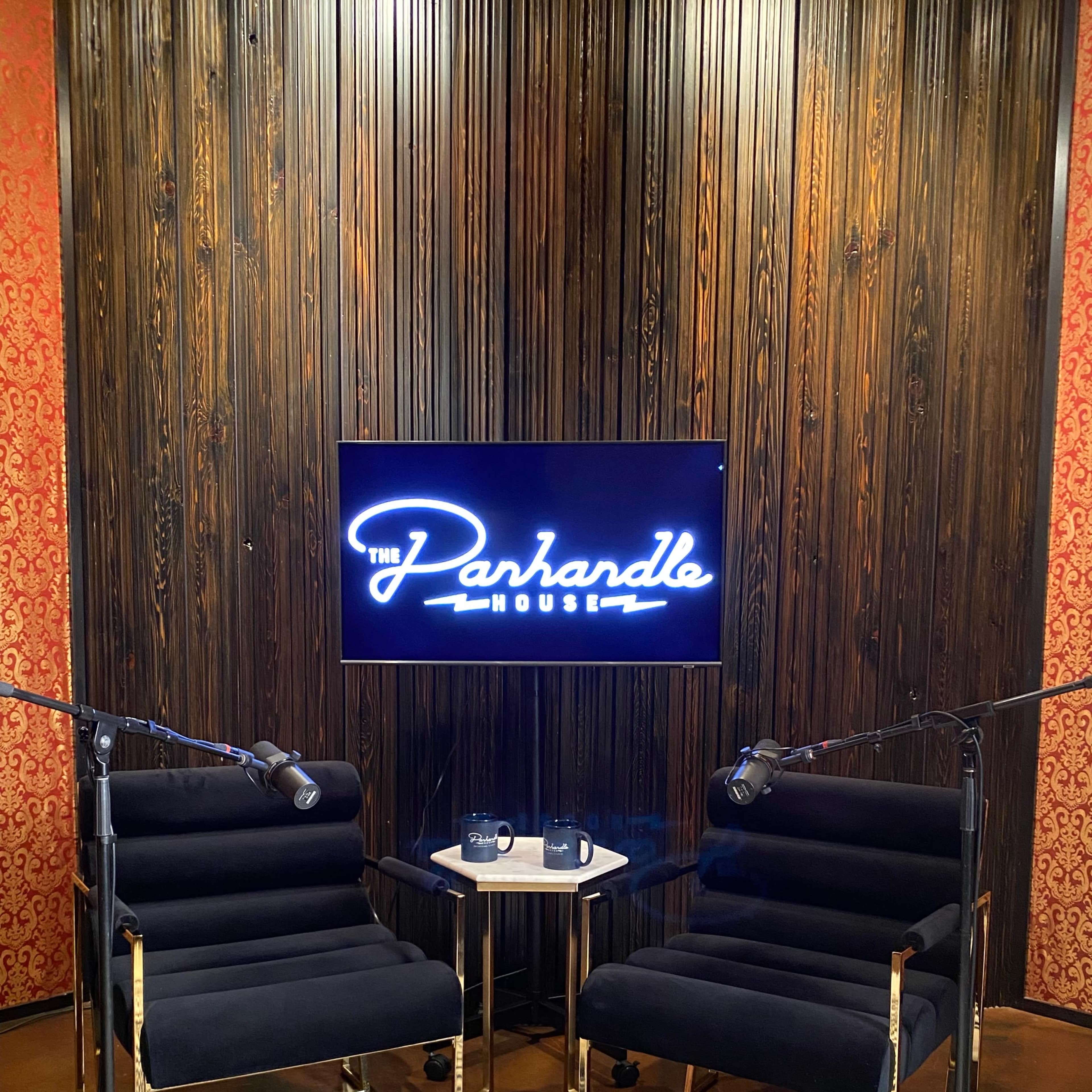 A podcast studio with two black chairs facing a small table, a television screen displaying "The Parhandle House," and microphones positioned for recording.