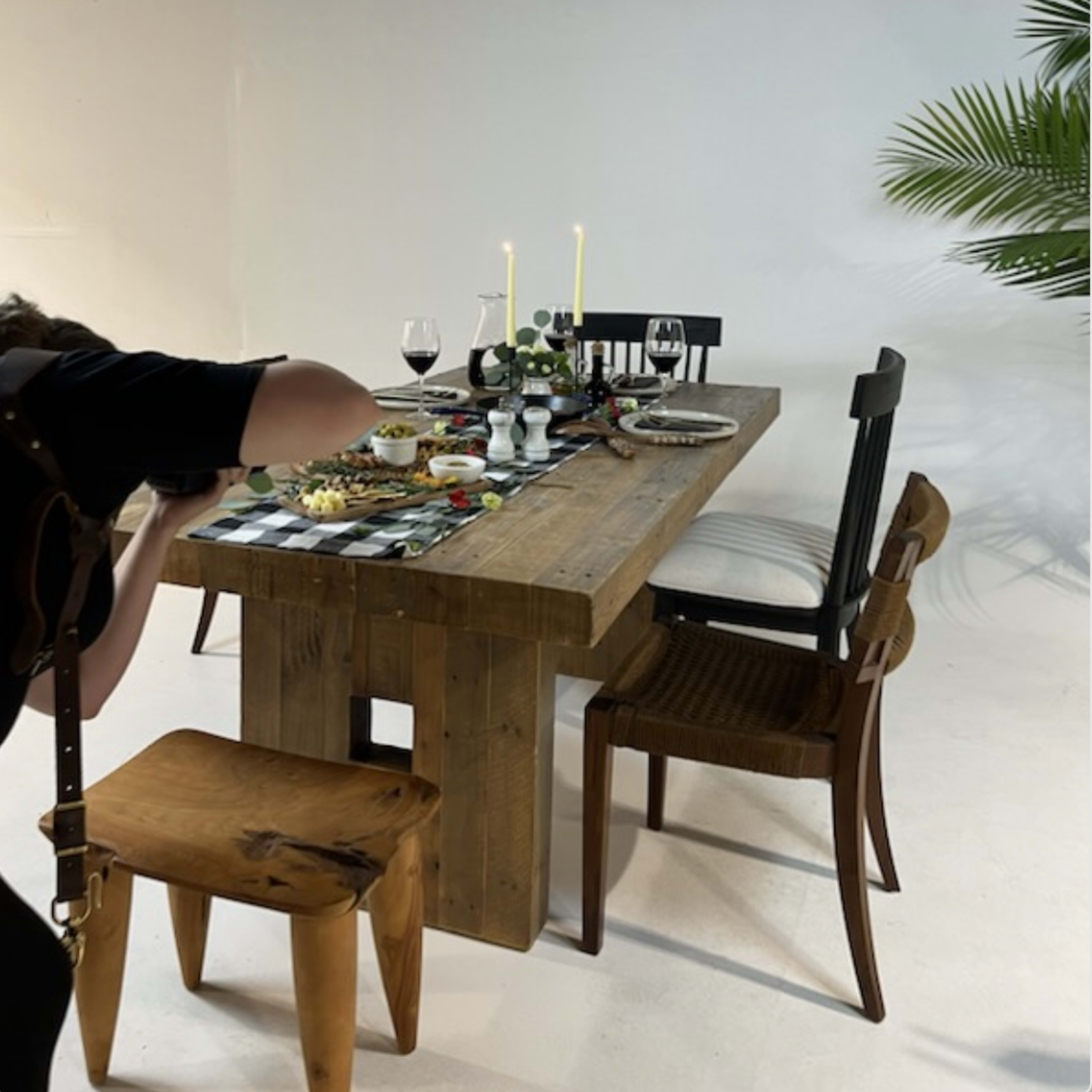 A wooden dining table is set with various dishes and two candles, surrounded by several chairs and a potted plant in the background.