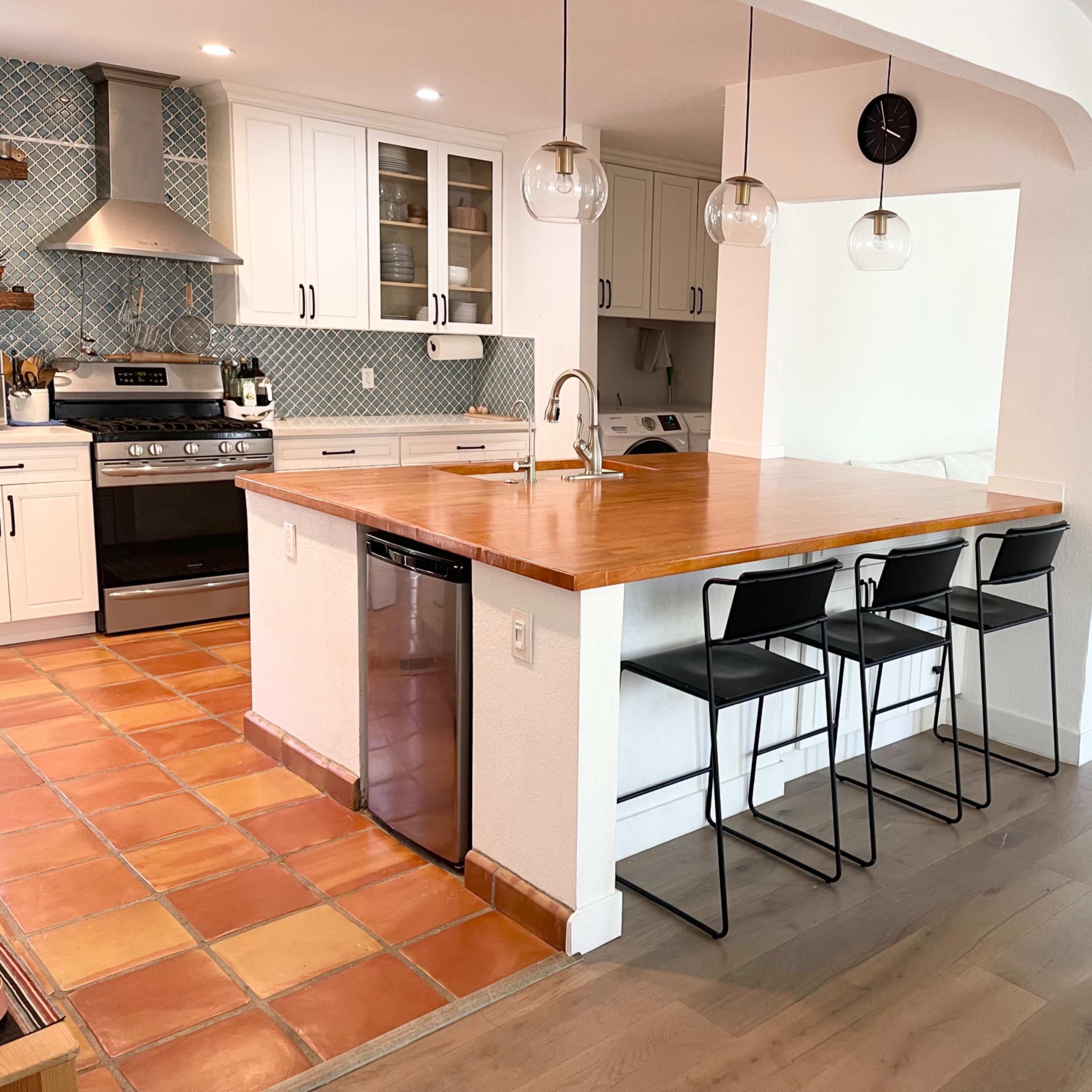 A modern kitchen with a wooden island, black bar stools, and a living area in the background featuring a white sofa and a coffee table.