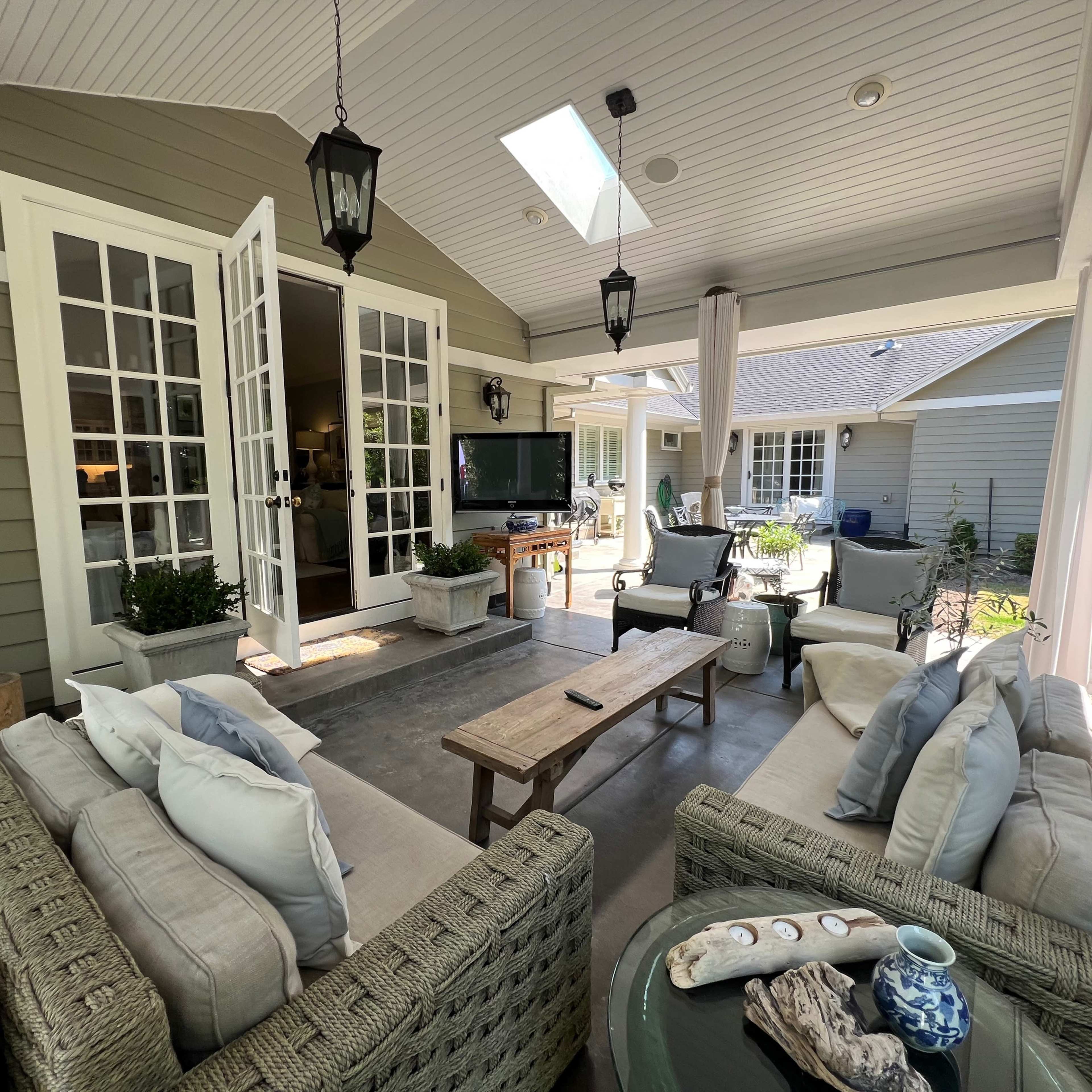 A covered outdoor living area with a sofa set, a coffee table, and a television mounted on the wall, surrounded by doors leading to other rooms.