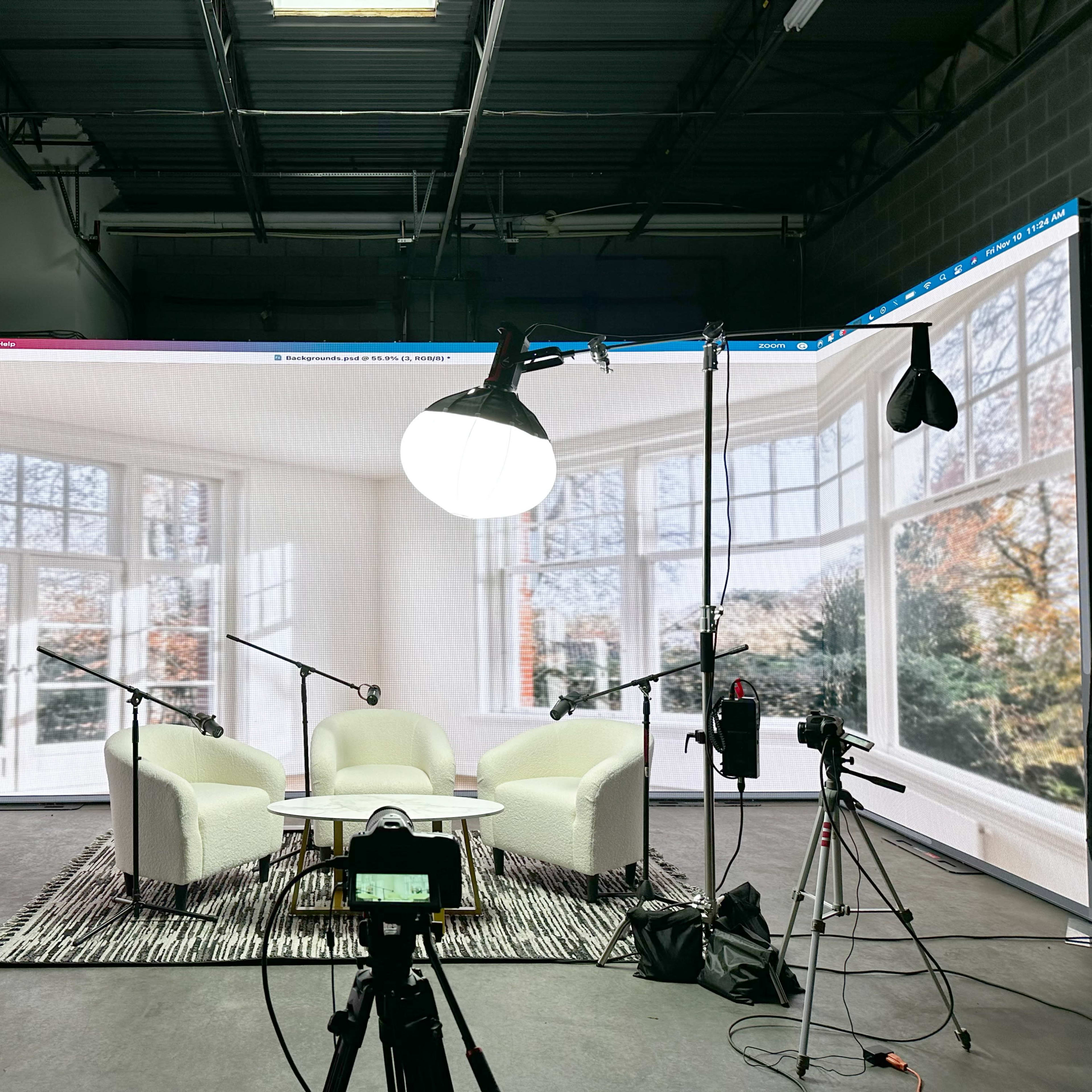 A studio setup with two white chairs and a circular table, surrounded by cameras and lighting equipment, against a backdrop of large windows displaying a garden view.