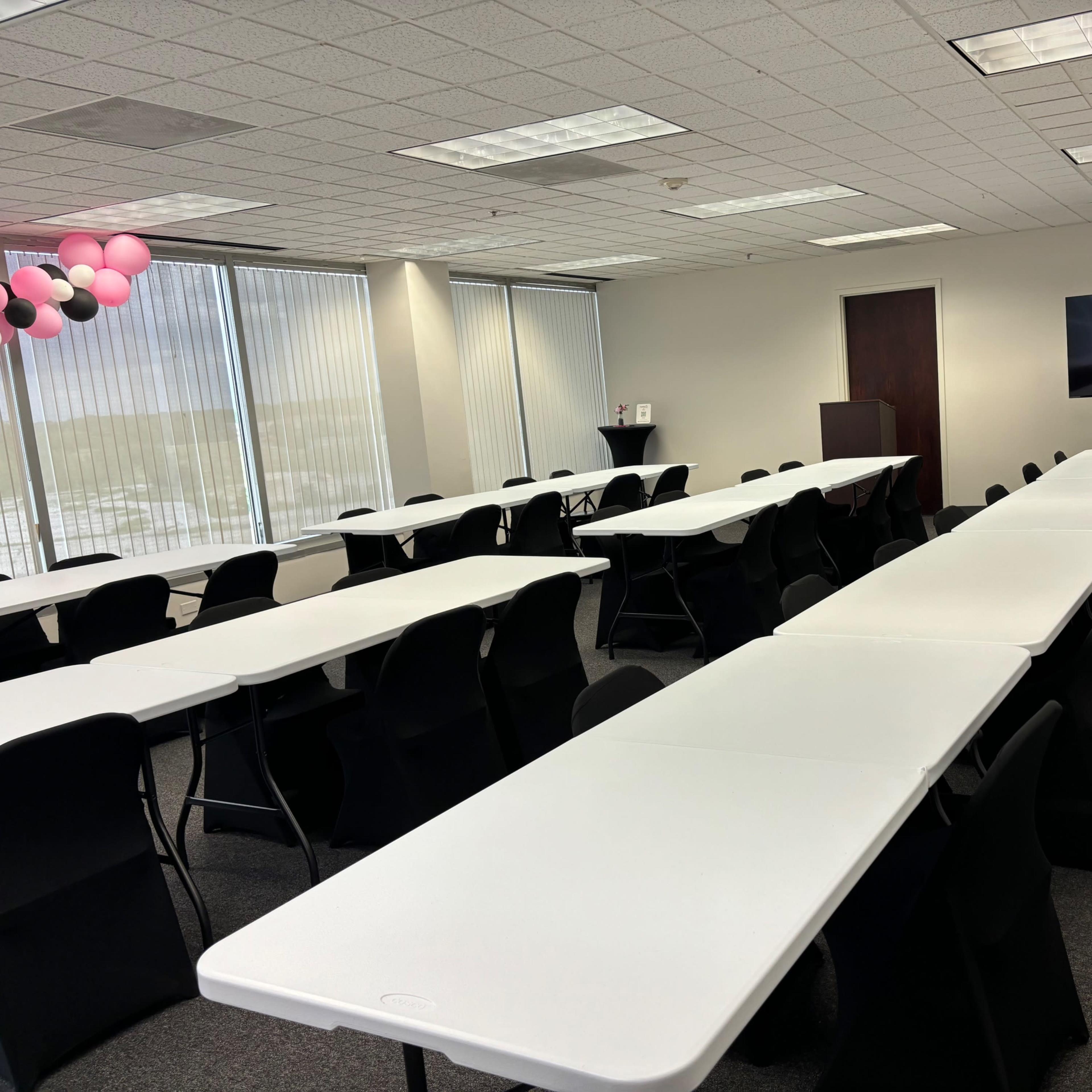 A conference room set up with multiple rows of white tables and black chairs, along with a large screen and decorative balloons.