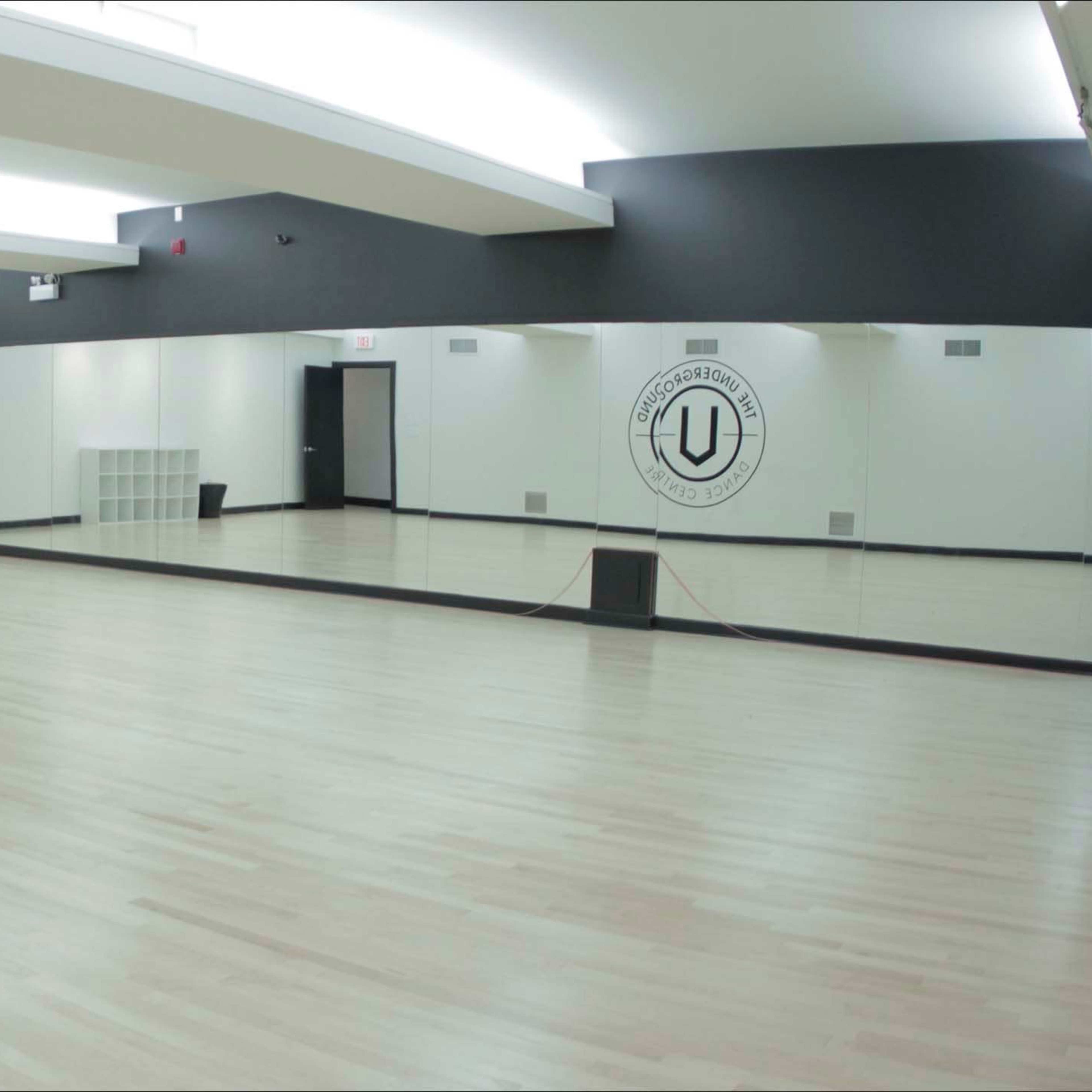 A spacious, empty dance studio with wooden flooring, large mirrors on one wall, and minimal storage units.