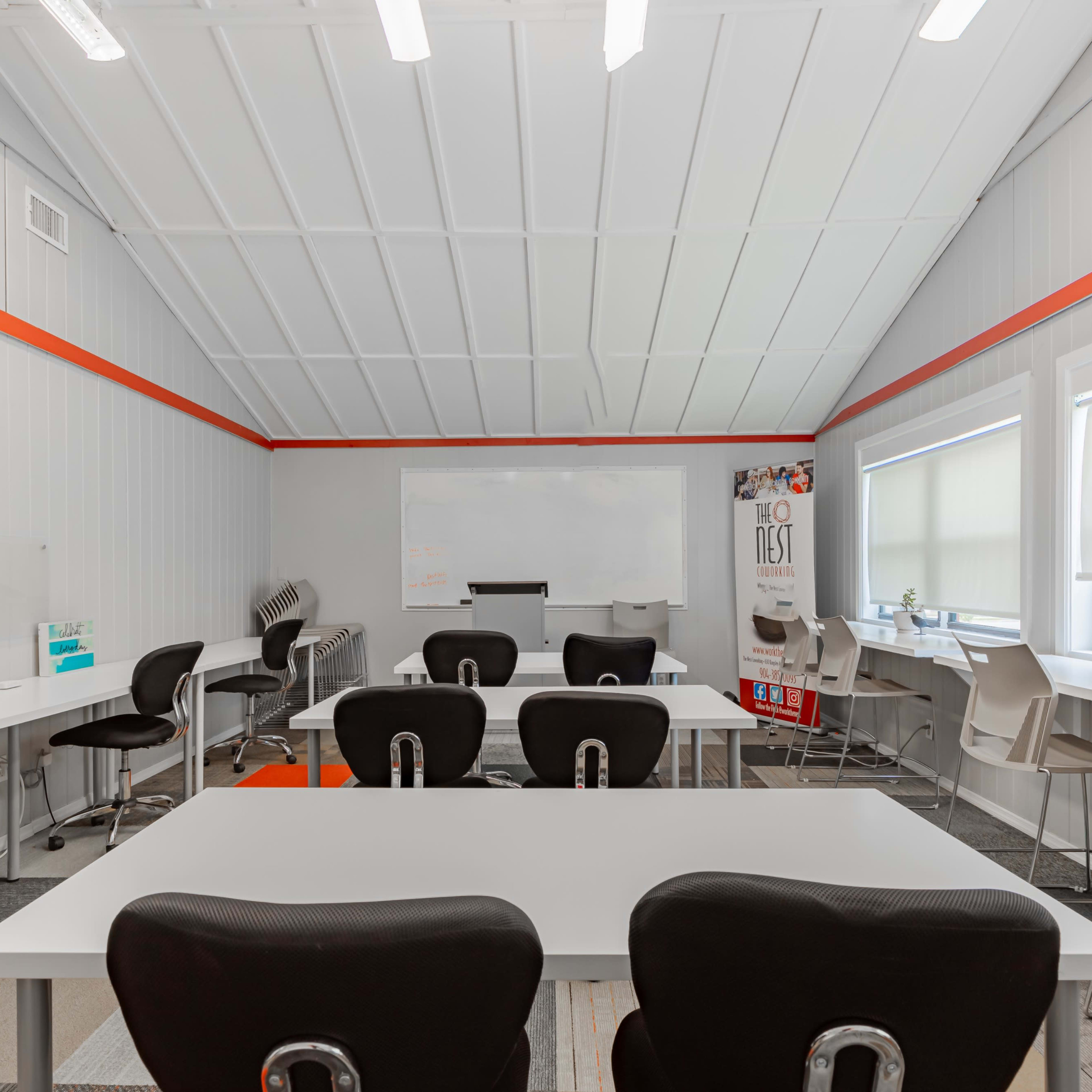 A modern classroom features white tables and black chairs arranged for group work, with large windows allowing natural light and a whiteboard at the front.