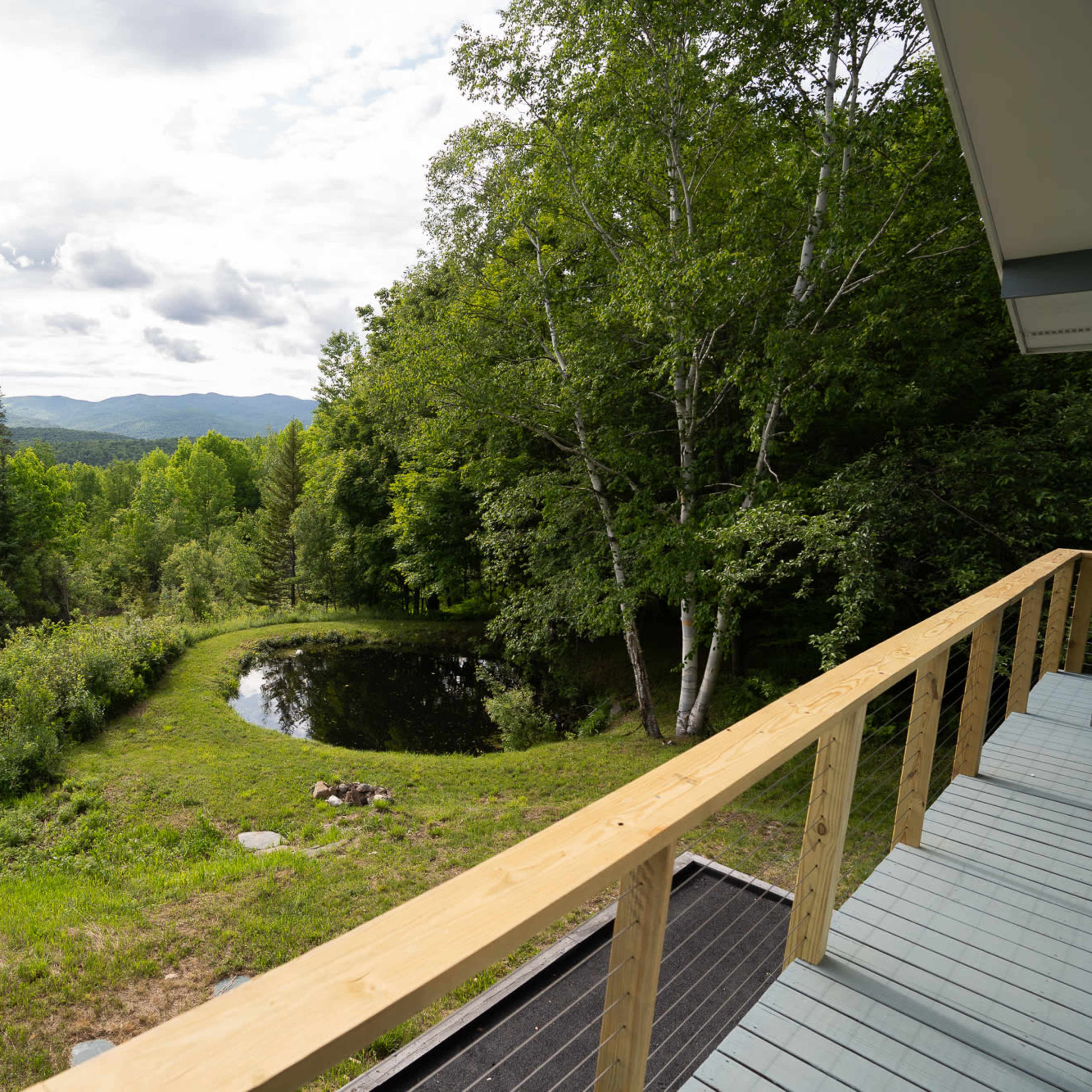 A wooden deck overlooking a pond surrounded by trees and mountains in the distance under a cloudy sky.