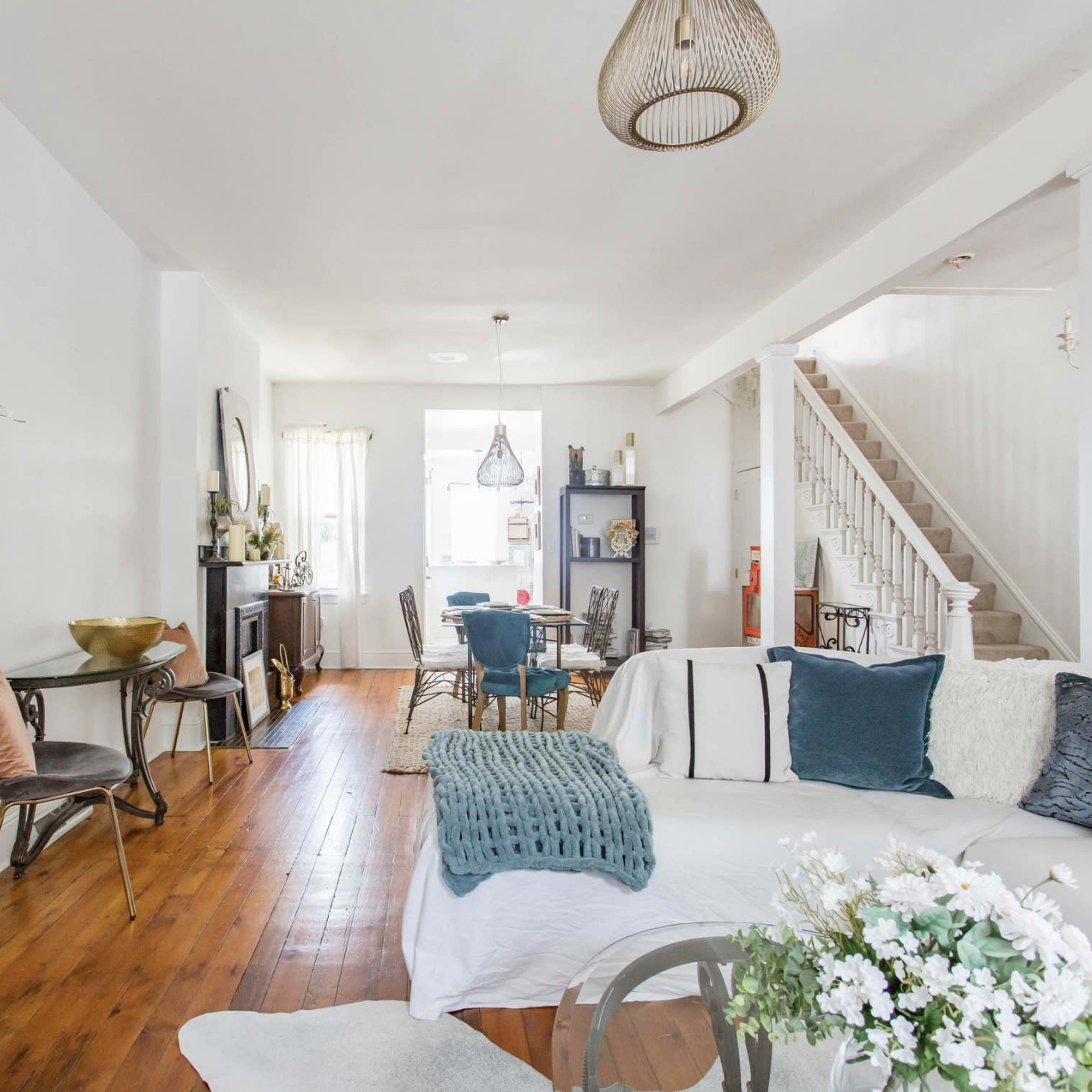 The living room features a white sofa, wooden flooring, and a staircase leading to an upper level, with various decorative elements and furniture throughout the space.