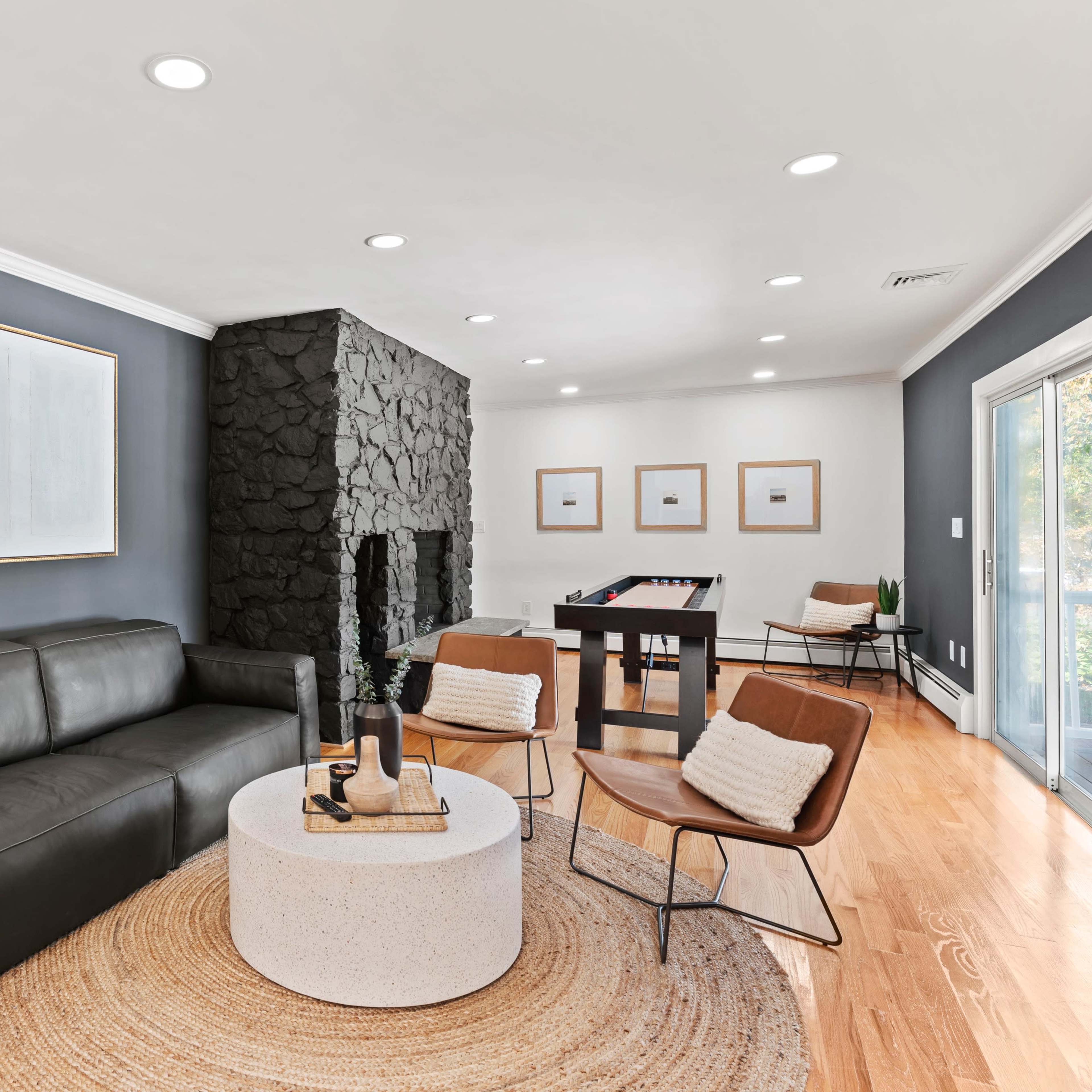 A modern living room featuring a dark sofa, a round coffee table, and a fireplace with a stone facade, complemented by a dining area in the background.