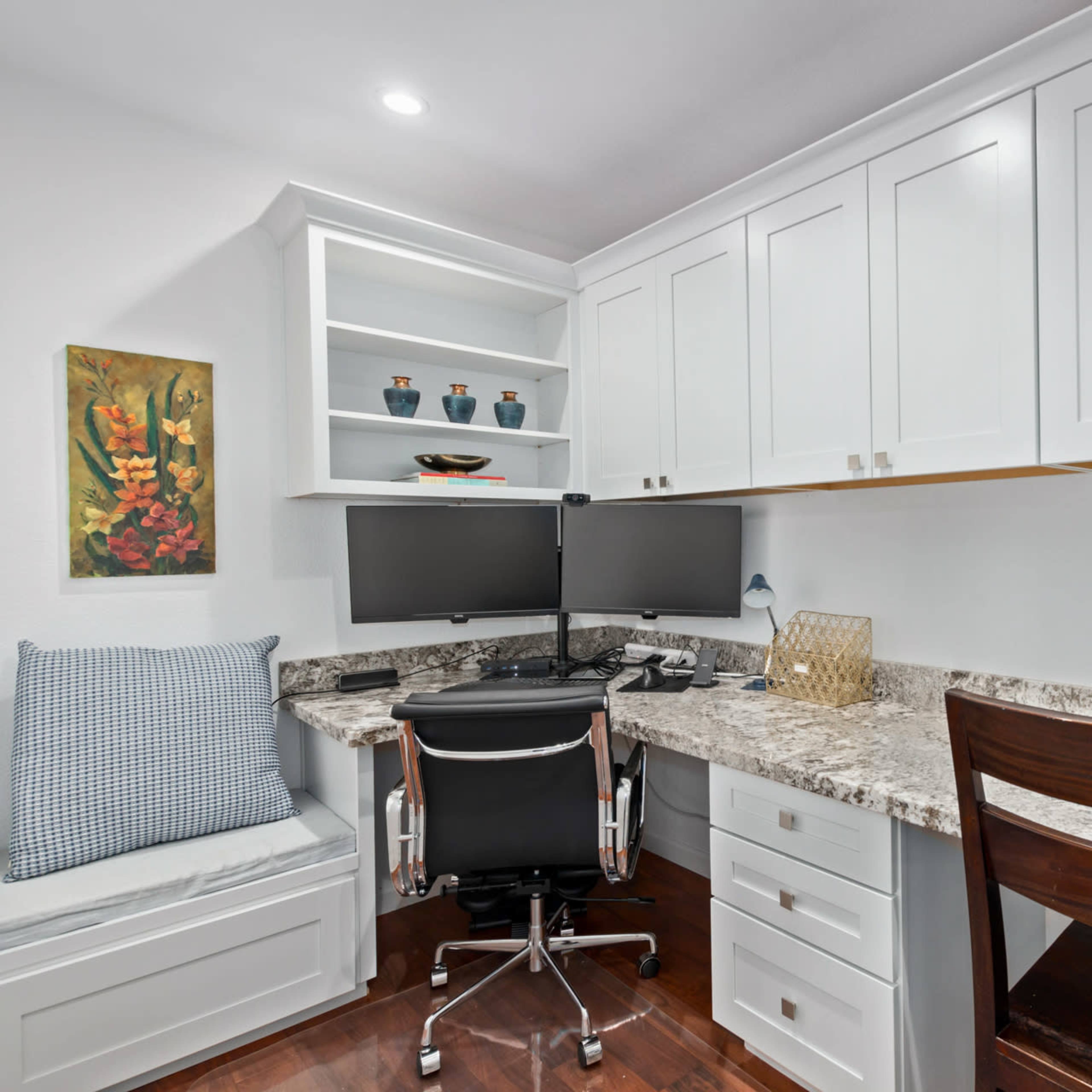 A compact home office features a granite countertop, two monitors, a swivel chair, and built-in cabinetry with decorative items.