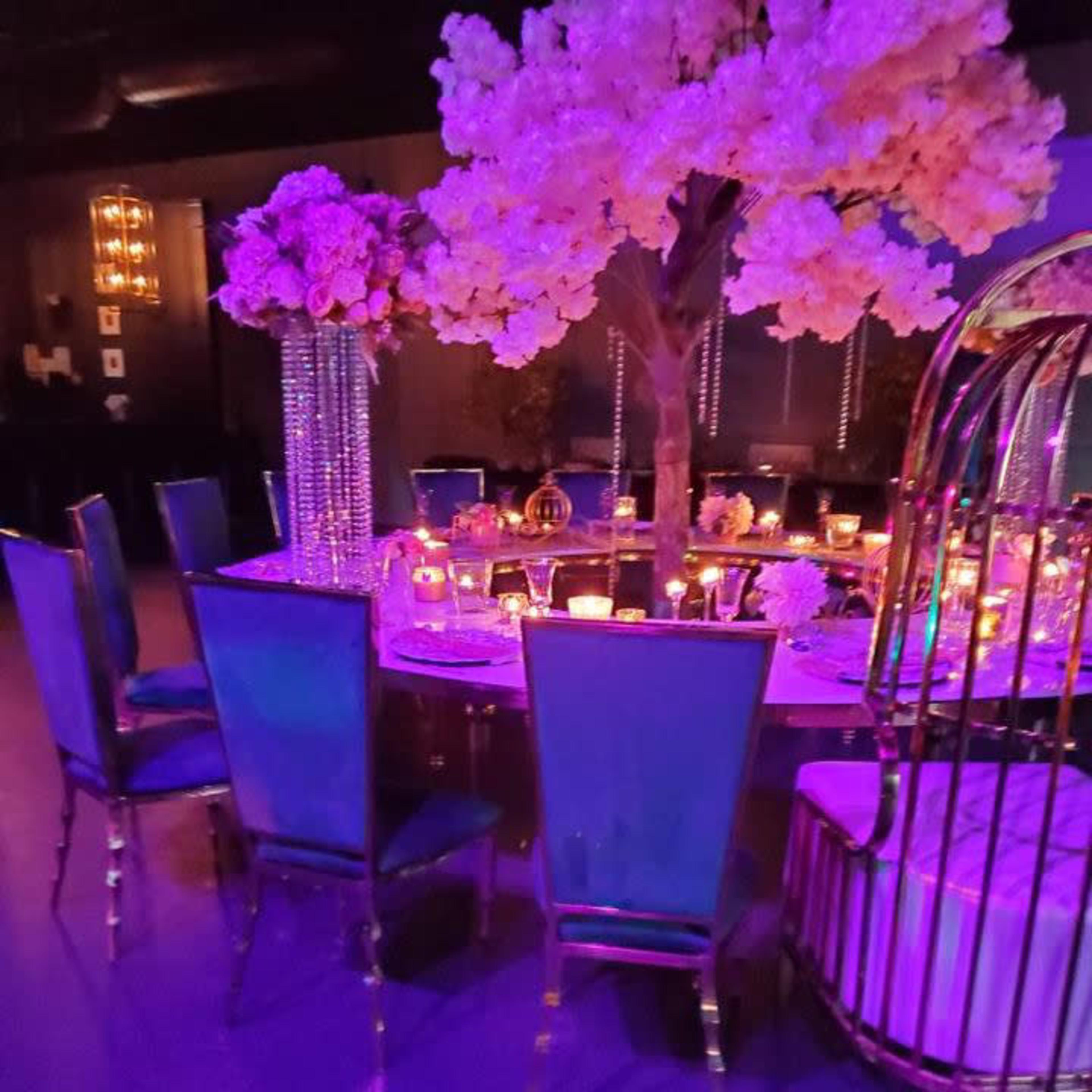 A dining table is elegantly set with candles and floral arrangements under a large cherry blossom tree, surrounded by blue chairs in a dimly lit space.