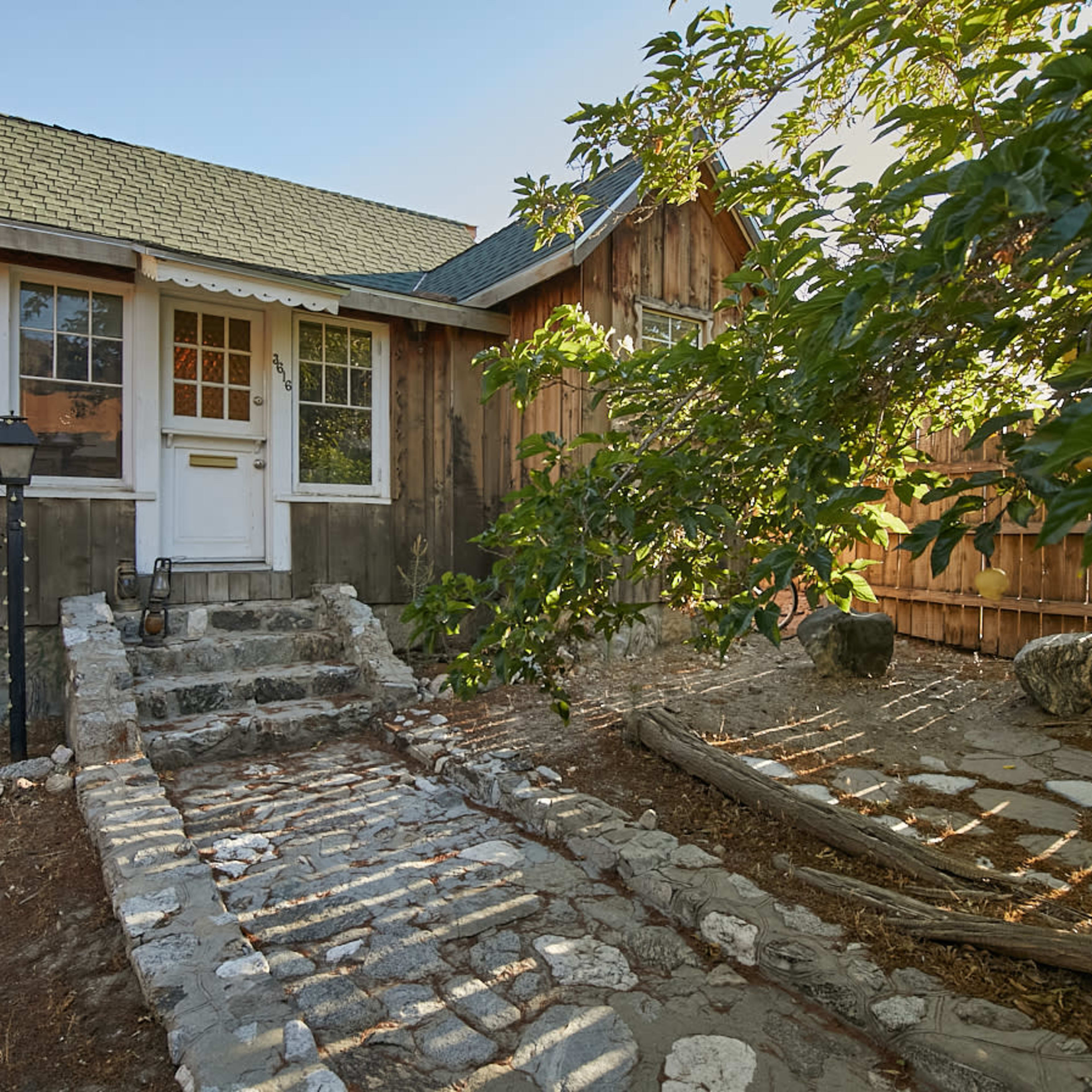 A stone pathway leads to the front door of a wooden house, surrounded by a fence and greenery.
