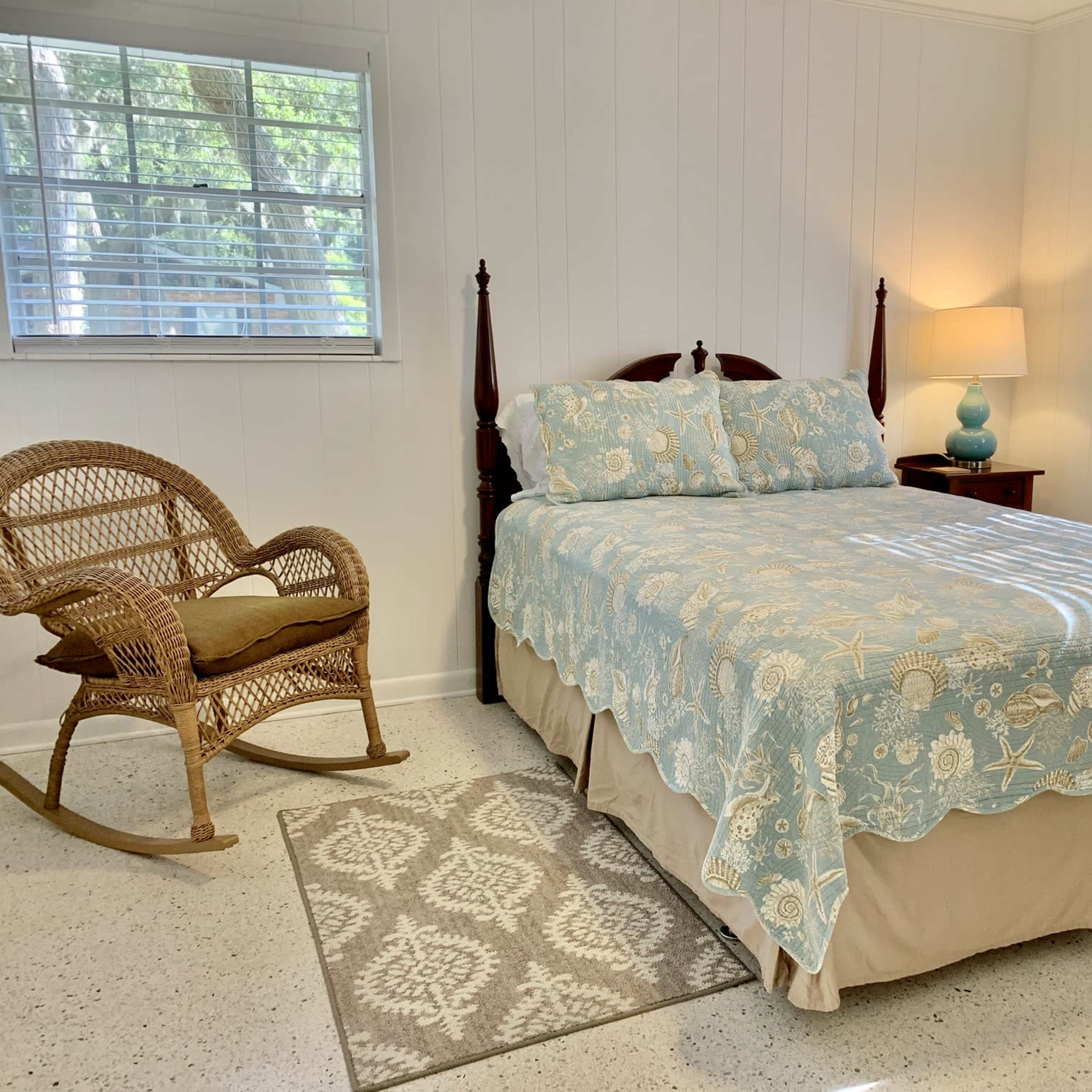 A bedroom with a bed covered in a patterned blue bedspread, a wicker chair, a bedside table with a lamp, and windows allowing natural light.