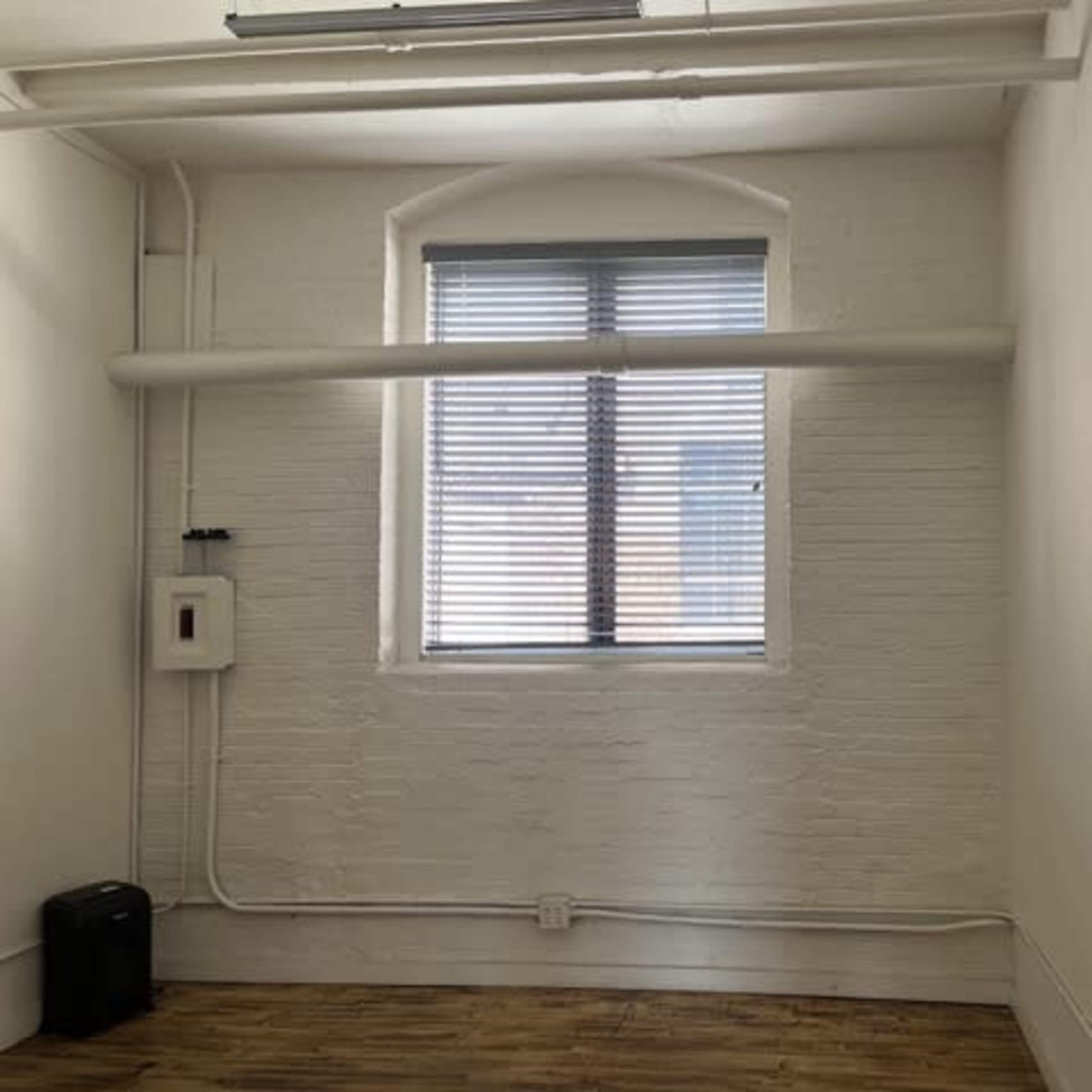 A small, empty room with a window covered by blinds, a wall with white brick, and a wooden floor.