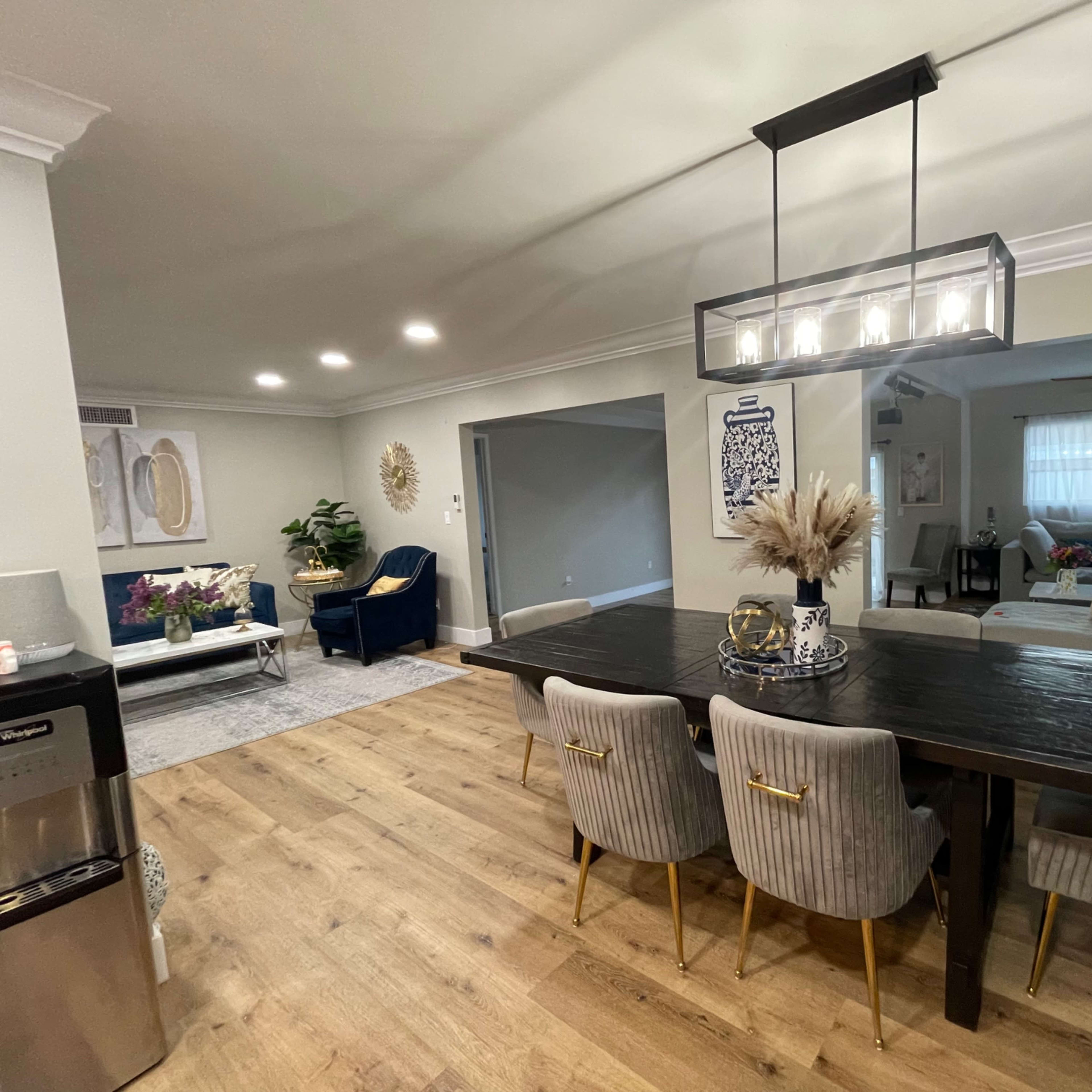 A modern dining area with a large wooden table surrounded by upholstered chairs, leading to a living room with a sectional sofa and a large window.