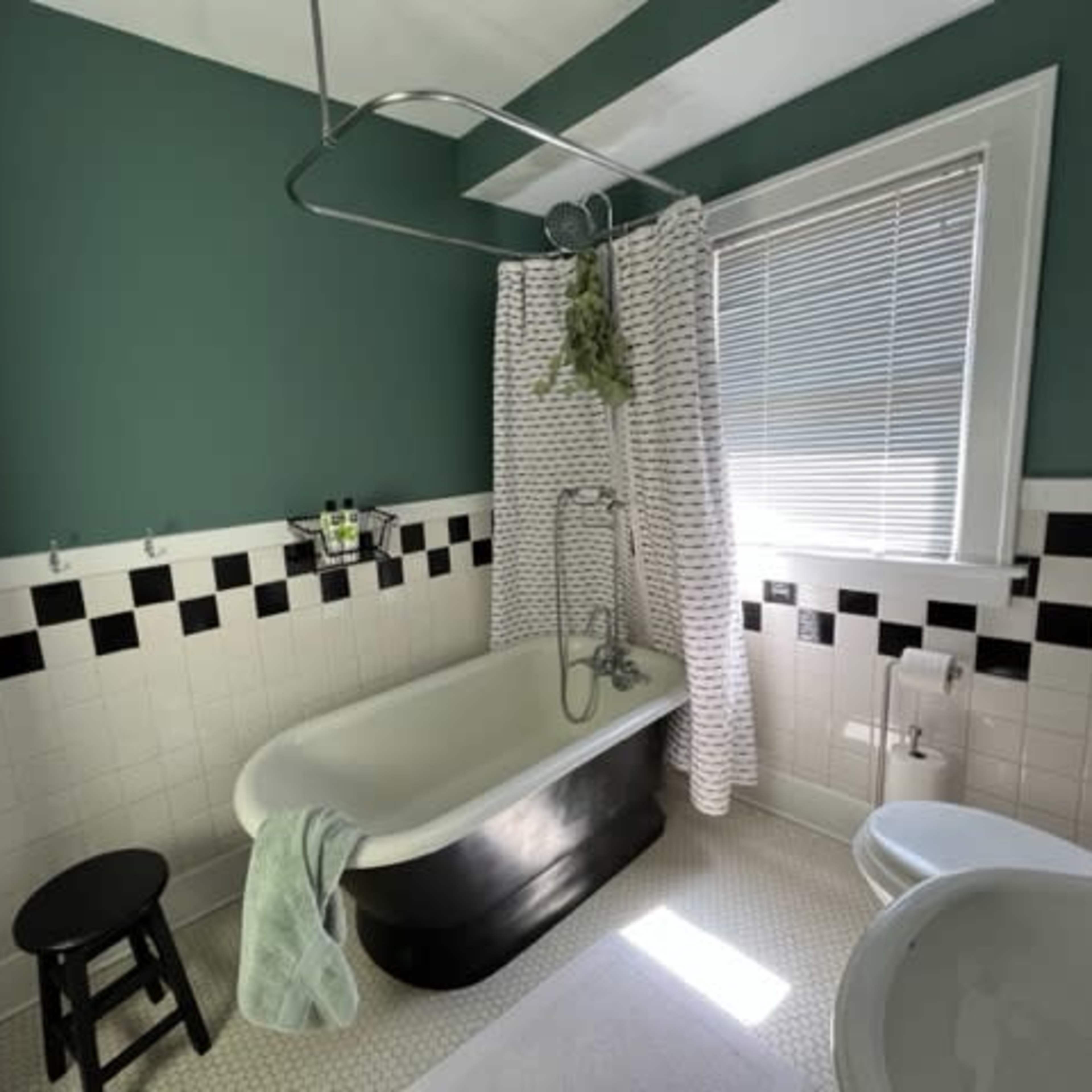 The bathroom features a clawfoot tub, a shower curtain, and black-and-white tile accents, with a small stool and a window allowing natural light.