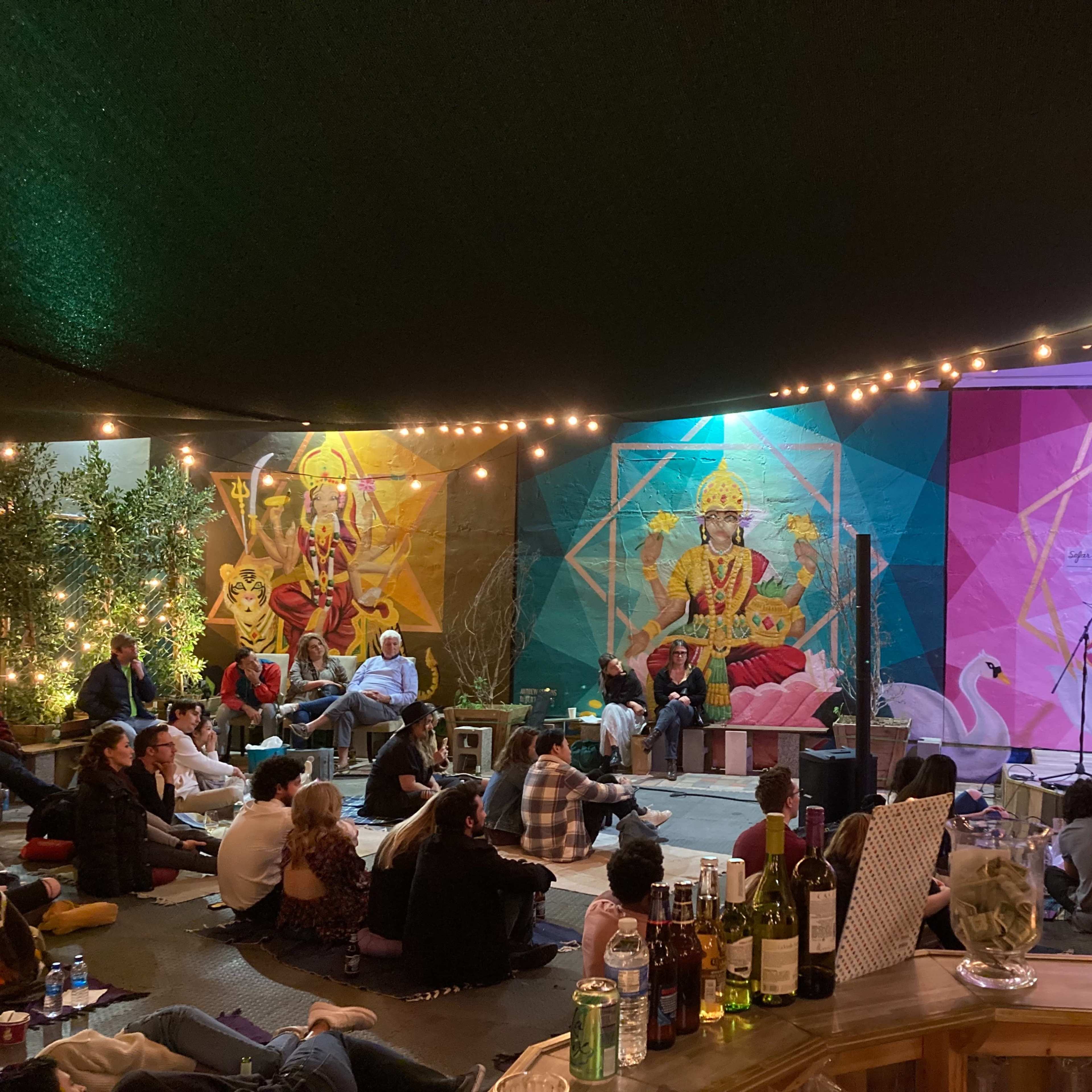 A musician performs on stage while an audience listens, surrounded by colorful murals and string lights in an outdoor space.