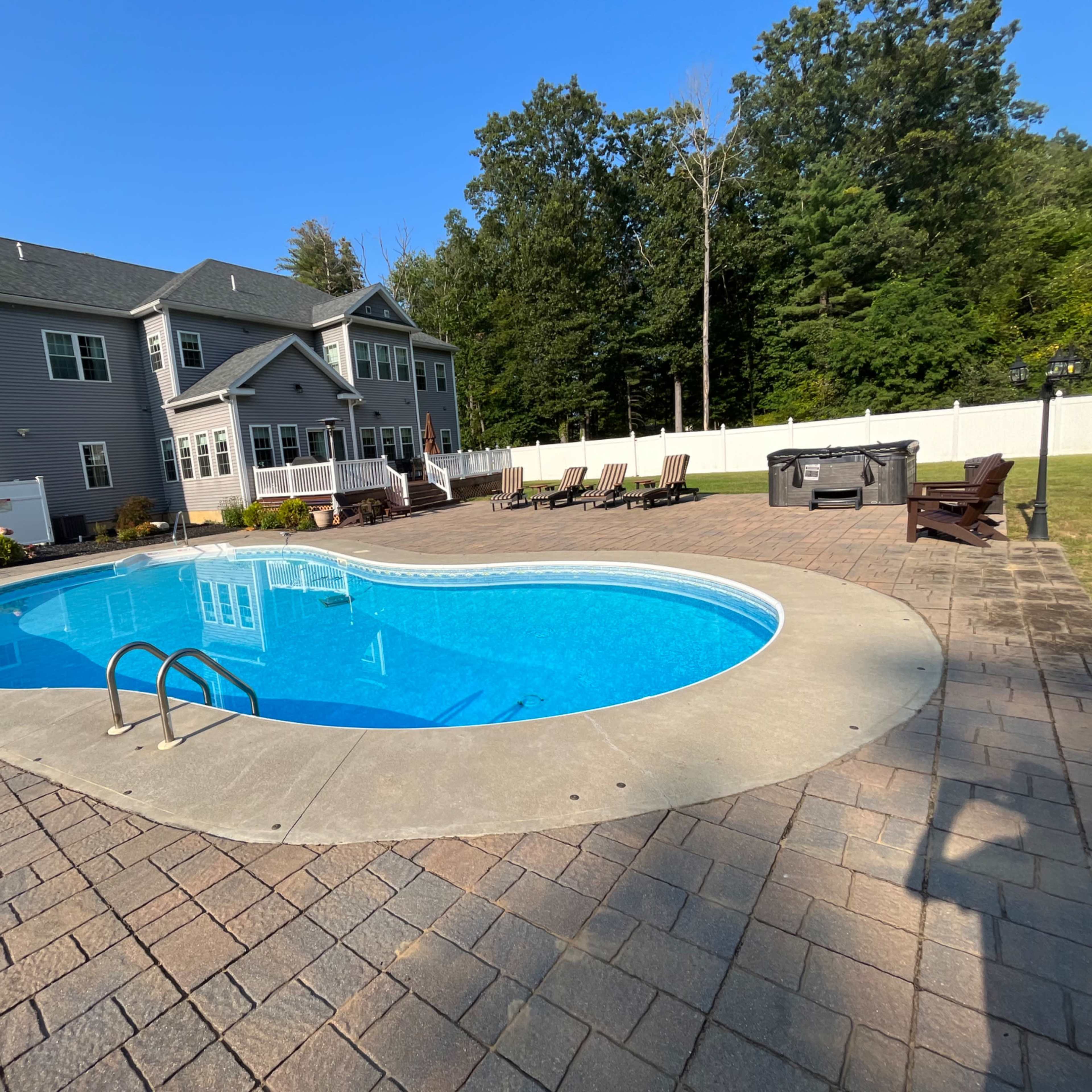 A residential backyard with a blue swimming pool surrounded by a stone patio and several lounge chairs, with a large house and trees in the background.