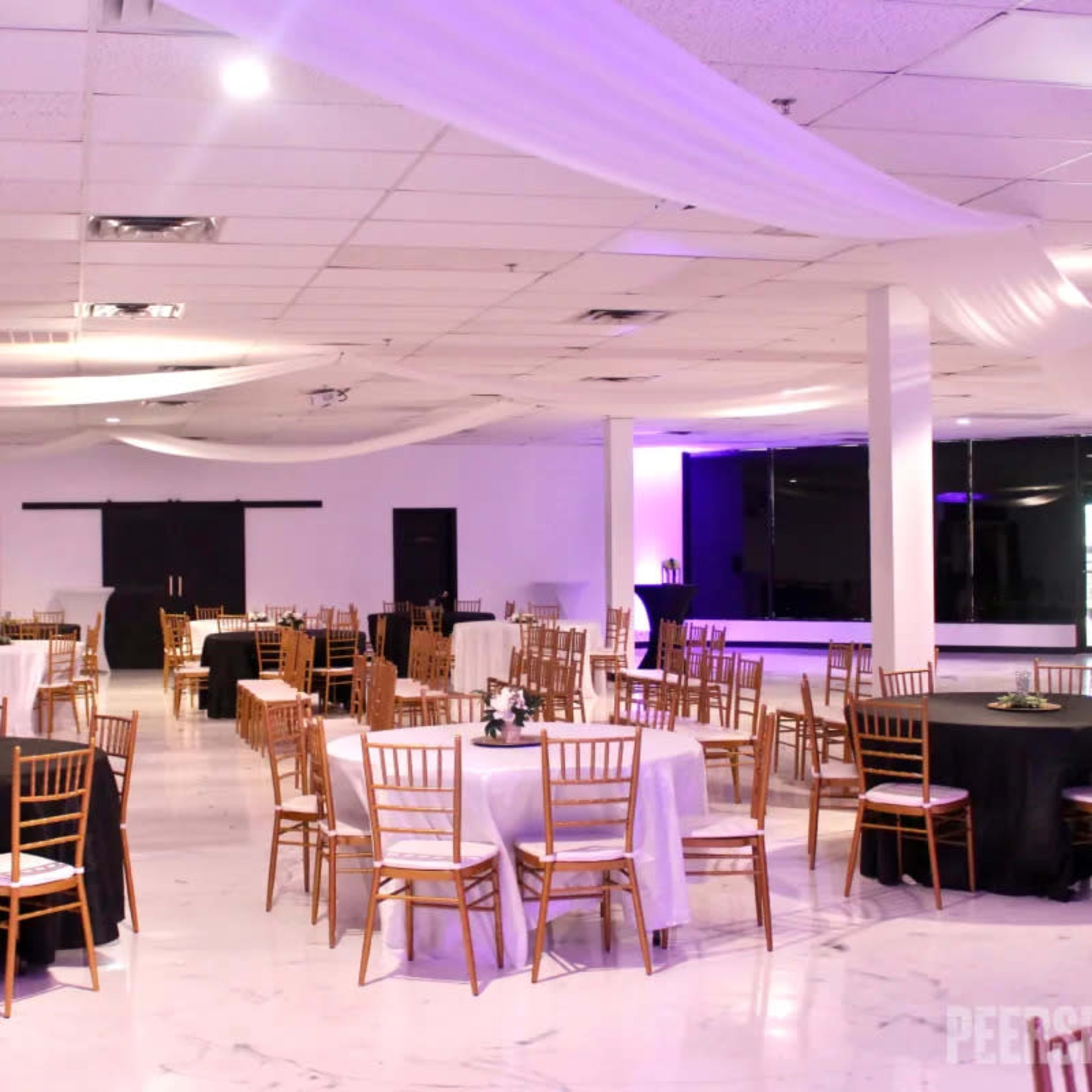 A spacious event venue with tables set in various arrangements, featuring black and white tablecloths, and draped fabric overhead.