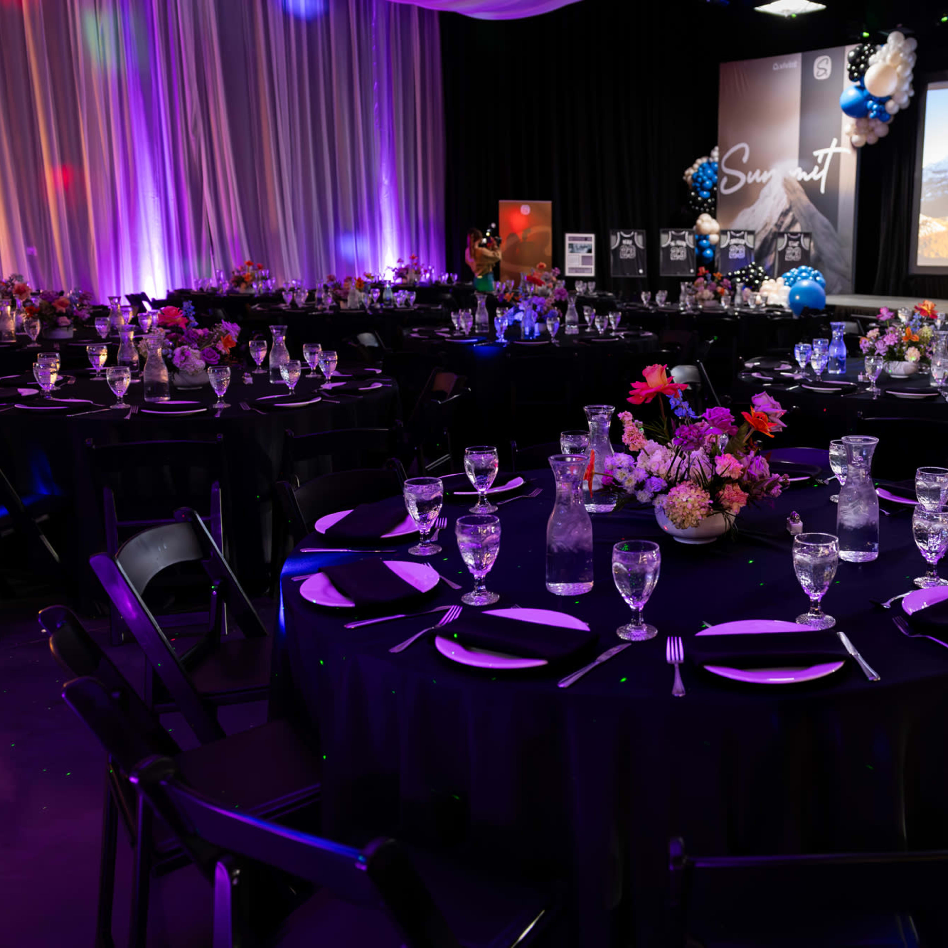 A banquet hall is set up with tables adorned with black tablecloths, centerpieces of flowers, and glassware, with colorful lighting and decorations in the background.